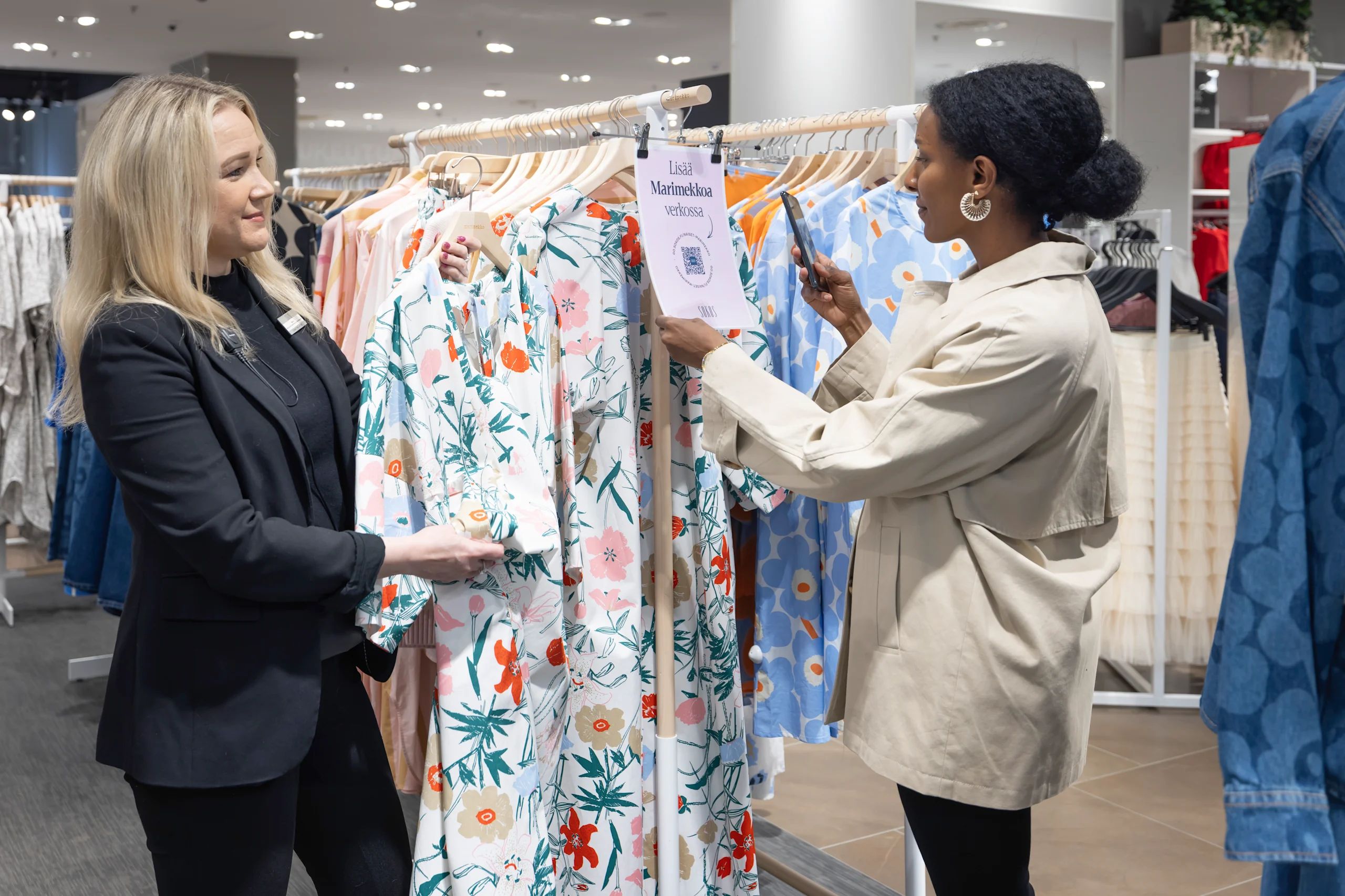 Myyjä esittelee asiakkaalle Marimekon mekkoja. Asiakas lukee kännykällä QR-koodia, jotta hän löytäisi saman mekon Sokoksen verkkokaupasta.