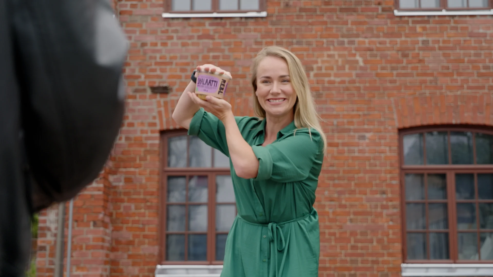 Elina pitelee Shalaatti-pakkausta käsissään ja hymyilee. Taustalla tiiliseinä.
