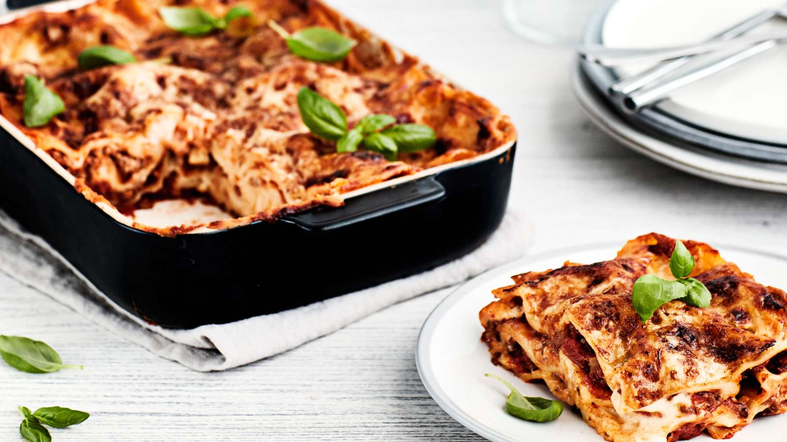 Lasagne mustassa uunivuoassa ja lautanen, jonka päällä basilikan lehdillä koristeltu annos lasagnea.