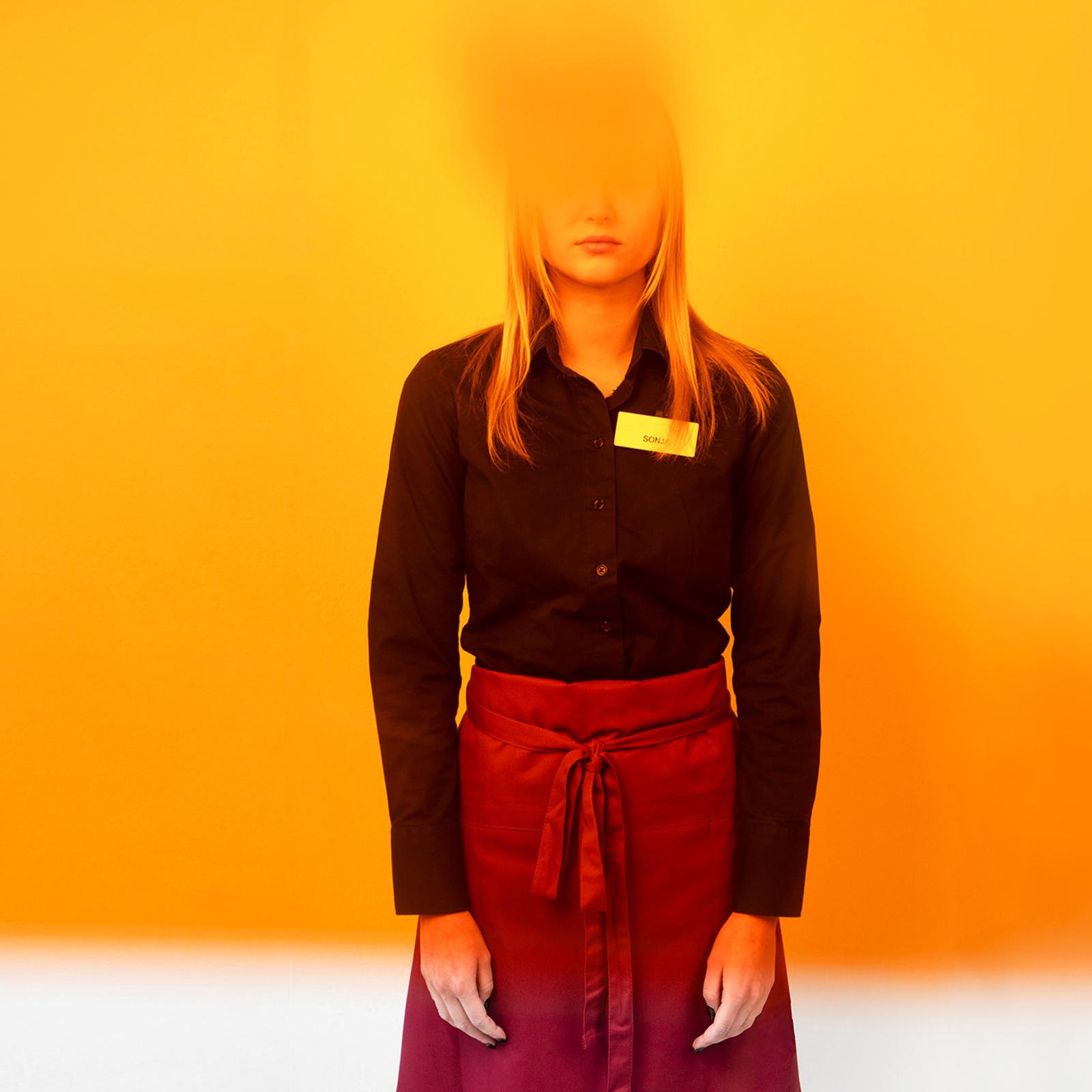 Essuun ja kauluspaitaan pukeutunut henkilö, jonka kasvot on sumennettu oranssilla värillä.