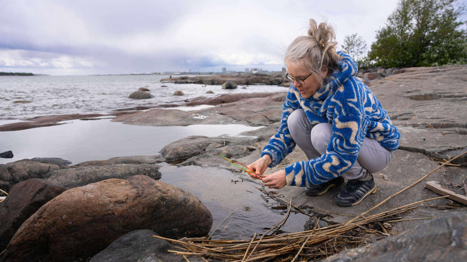 Kuvataitelija Tuula Närhinen on kyykistyneenä meren rannan kalliolla. Kädessään hänellä on rantaan huuhtoutunutta muovia.