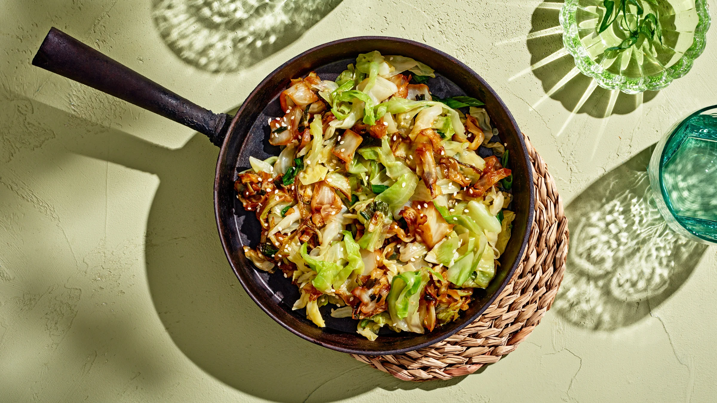 Paistettu varhaiskaali valurautaisella pannulla. Pannun alla pannunalunen ja vieressä vesilaseja.