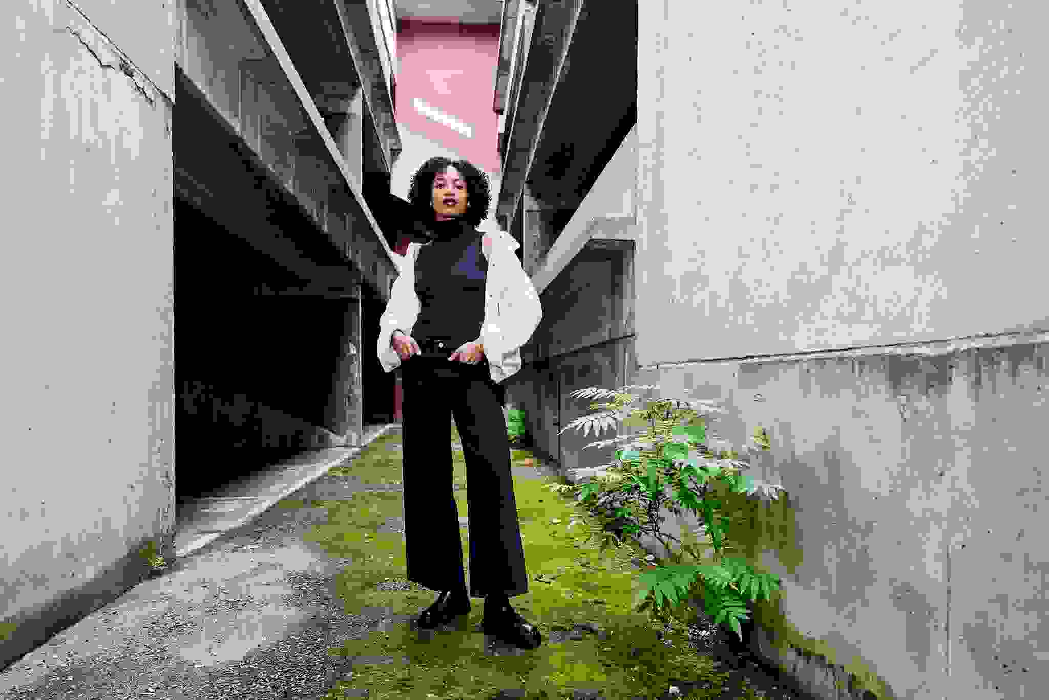 Nainen seisoo WKLY.-merkin vaatteet päällä betonisessa kaupunkimiljöössä