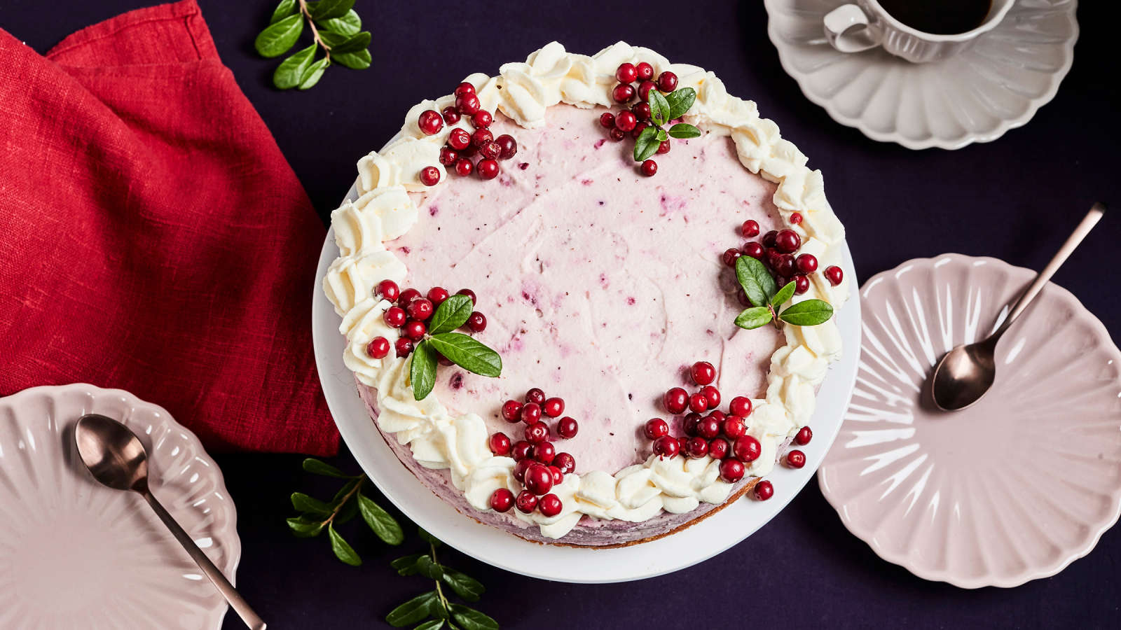 Puolukkamoussekakku, jonka vieressä kaksi kakkulautasta, kahvikuppi ja punainen liina. 