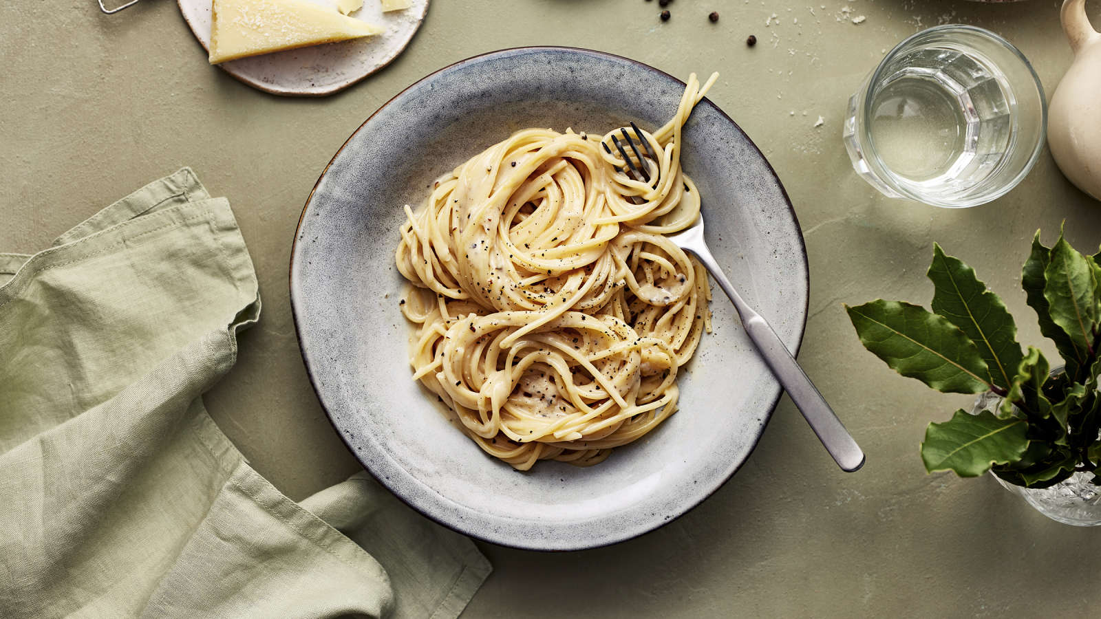 Cacio e pepe -pastaa lautasella. Annoksen vieressä vesilasi, vihreä liina, pecorinojuustoa, pippureita ja kasvin lehtiä.