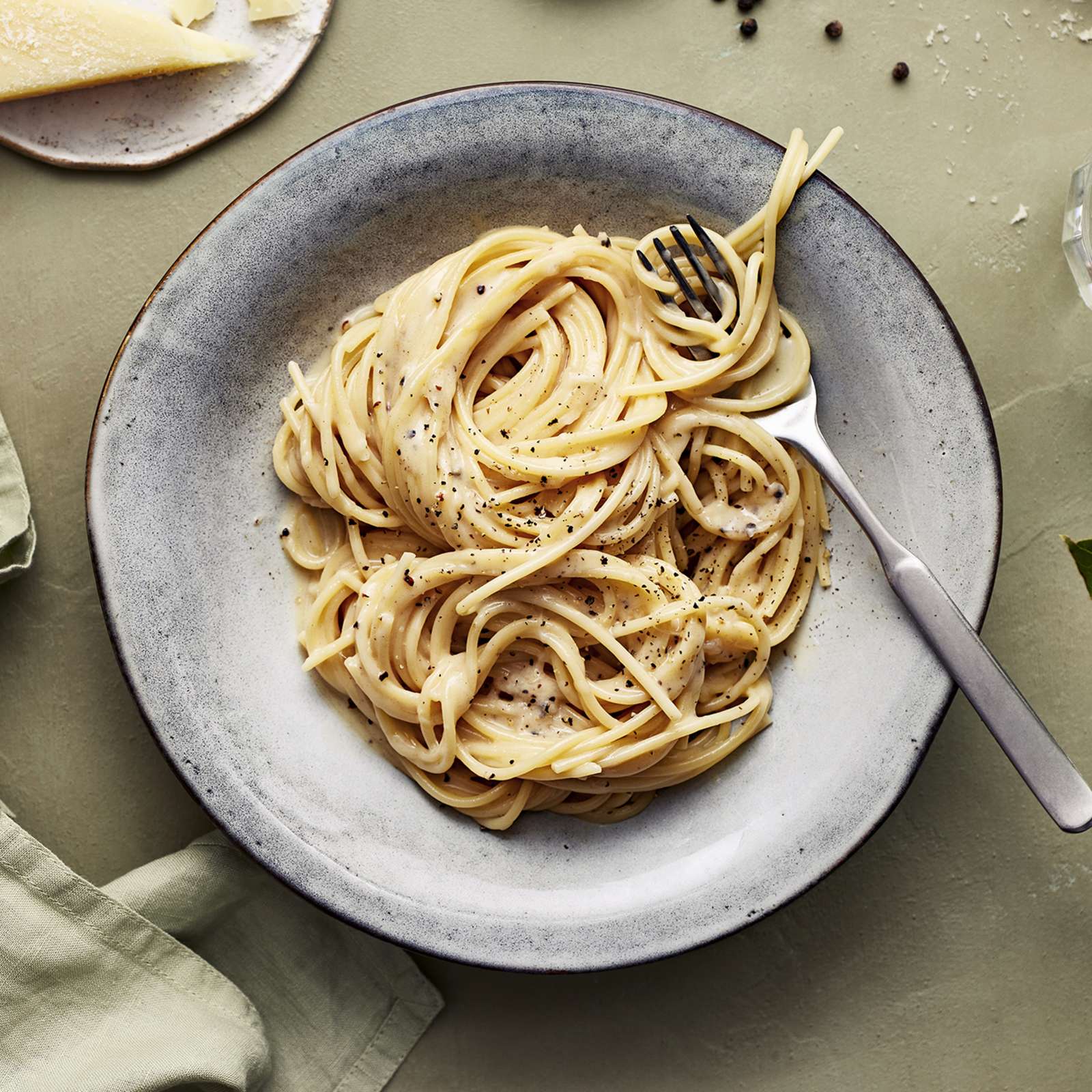 Cacio e pepe -pastaa lautasella. Annoksen vieressä vesilasi, vihreä liina, pecorinojuustoa, pippureita ja kasvin lehtiä.