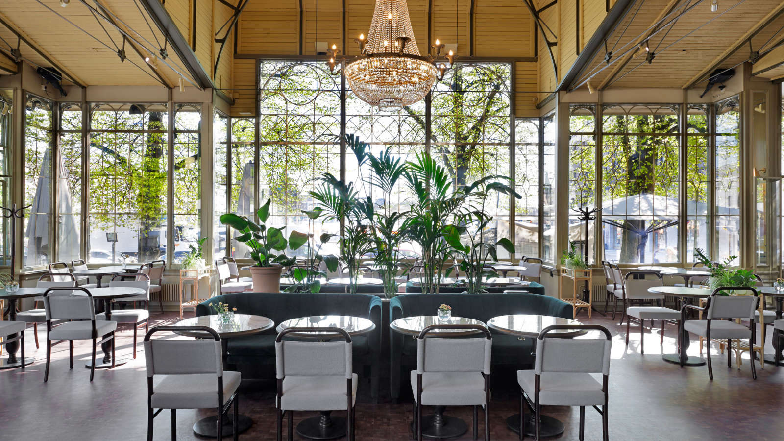 Kappelin kahvilasali, jossa on pöytiä ja tuoleja, suuri kristallikattovalaisin ja ikkunoista näkymä vehreään Esplanadin puistoon