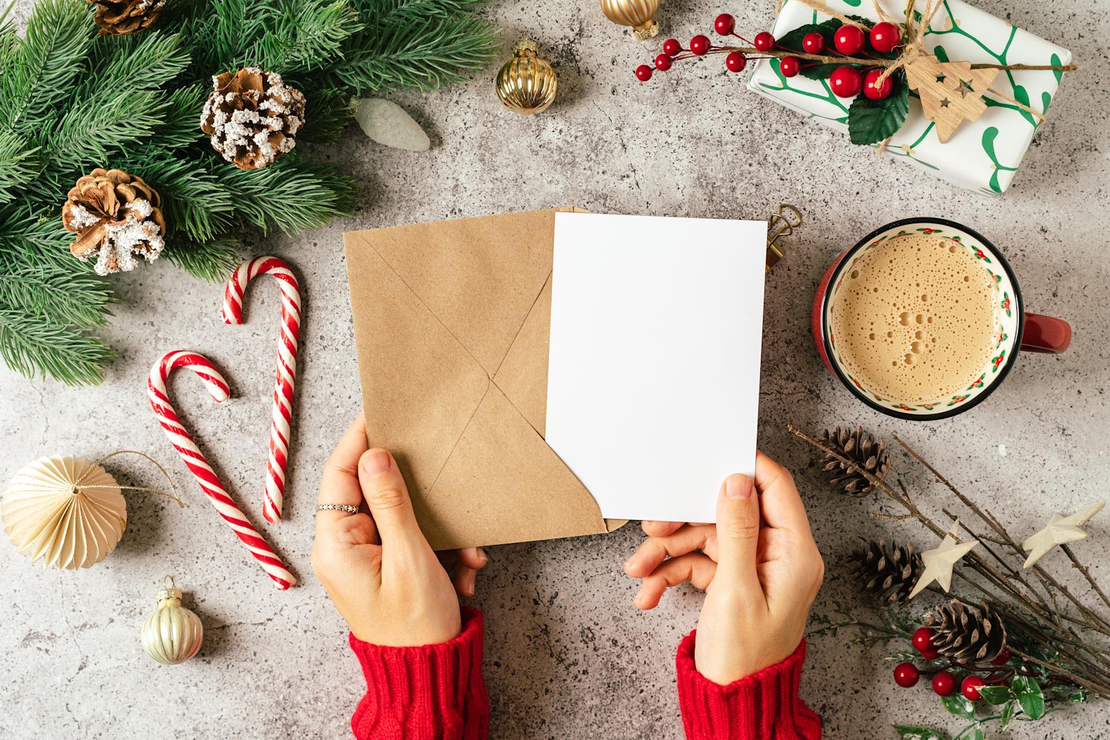Ylhäältä päin kuvatut kädet laittaa kirjekuoreen korttia, pöydällä joulukoristeita