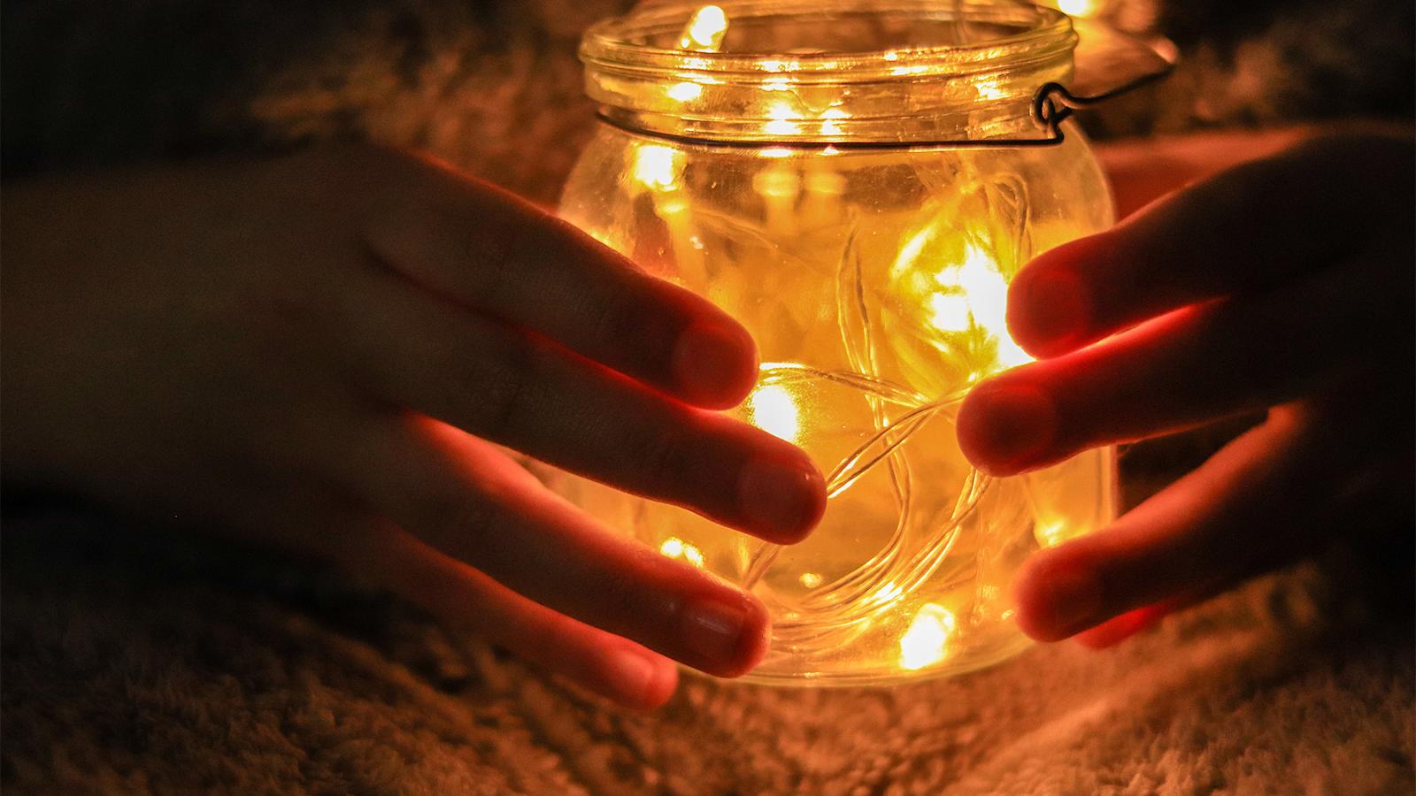 Kädet valaistun lasipurkin ympärillä.