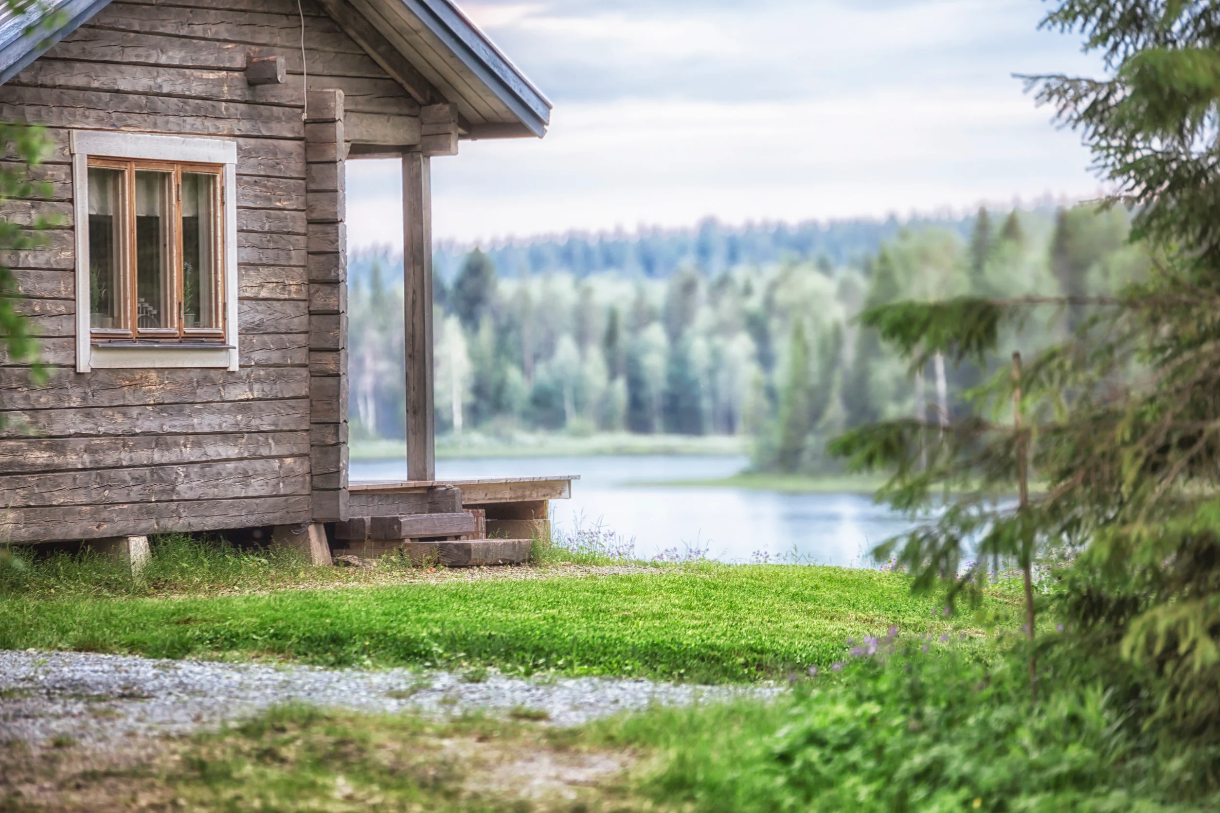 Hirsimökki järven rannalla. Mökin puupinta on kulunut. Sen edustalla on kuisti.