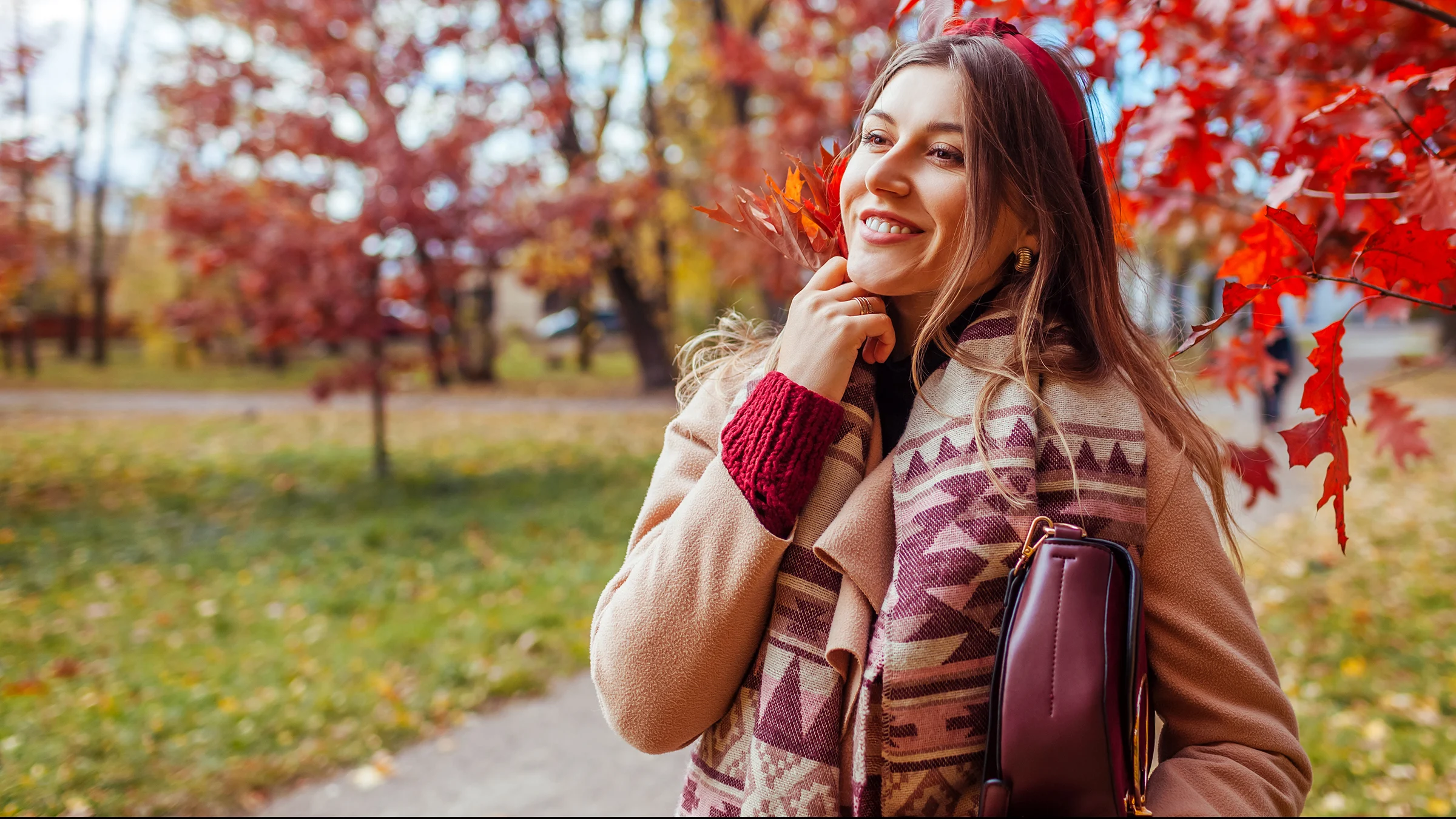 Nainen syksyisessä puistossa päällään beige takki ja viininpunainen neule ja laukku.
