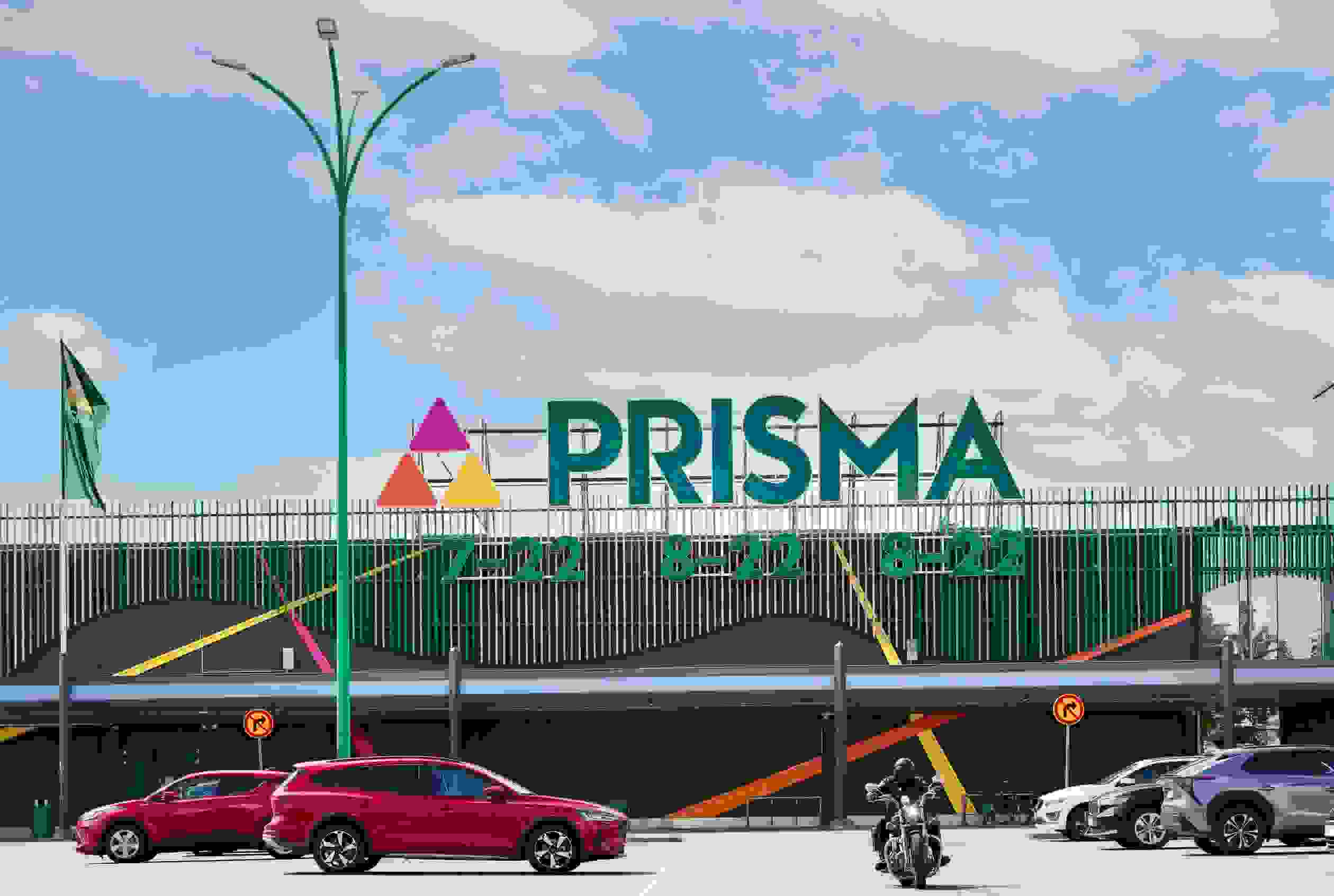 Prisman seinä ja kyltti näkyy parkkipaikalla autojen takana, moottoripyöräilijä ajaa parkkipaikalla kuvaajaa kohti. Yllä näkyy sininen taivas ja valkoisia pilviä.