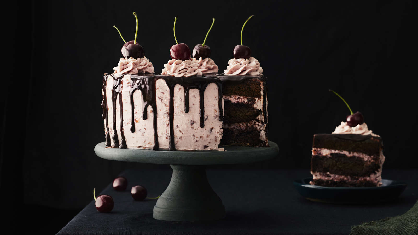 Tuoreilla kirsikoilla, pursotuksilla ja sulatetulla suklaalla koristeltu suklaa-kirsikkakakku tummalla, jalallisella kakkuvadilla. Kakusta leikattu pala lautaselle.