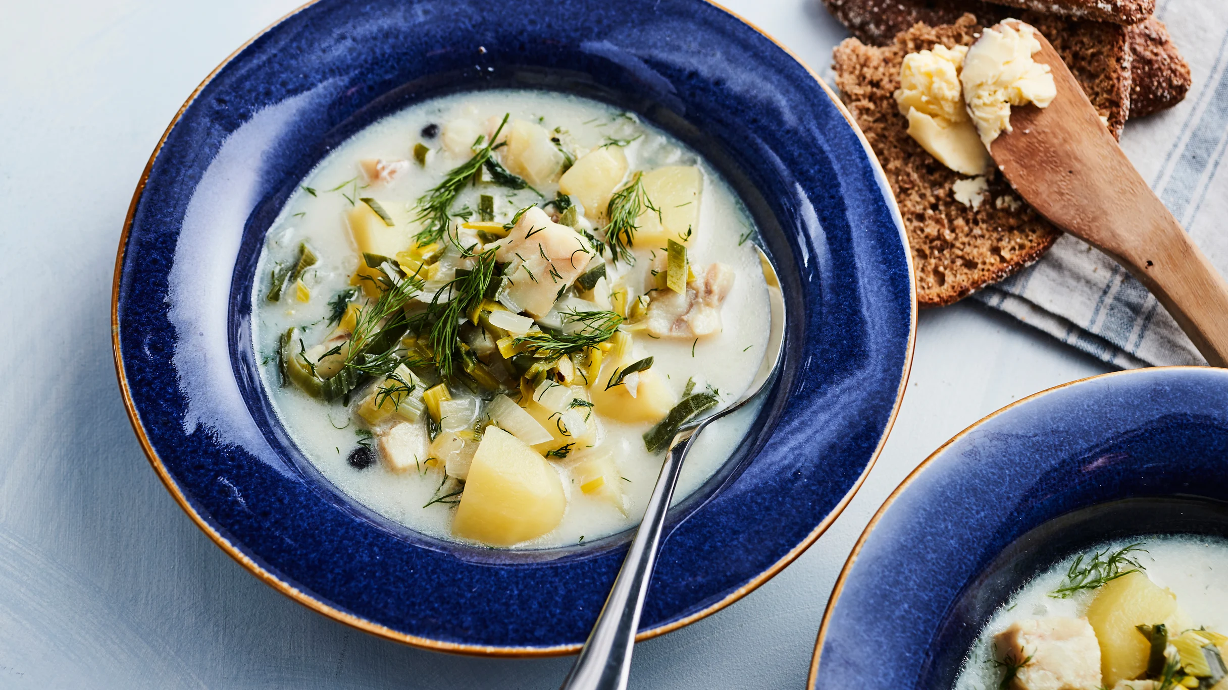 Sinisellä lautasella annos kalakeittoa ja lusikka. Vieressä voideltu ruisleipä, puinen leipäveitsi ja toinen annos keittoa.