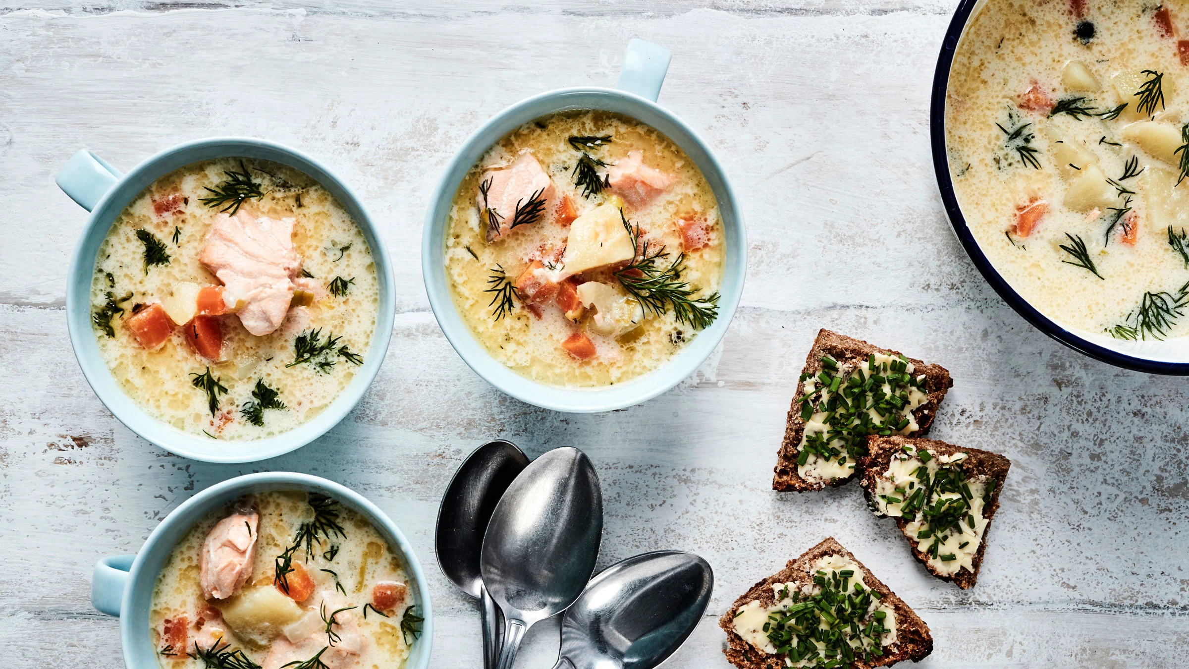 Kolme lohikeittoannosta, keittokattila, lusikoita ja persiljasilpulla koristeltuja voideltuja ruisleipäpaloja.