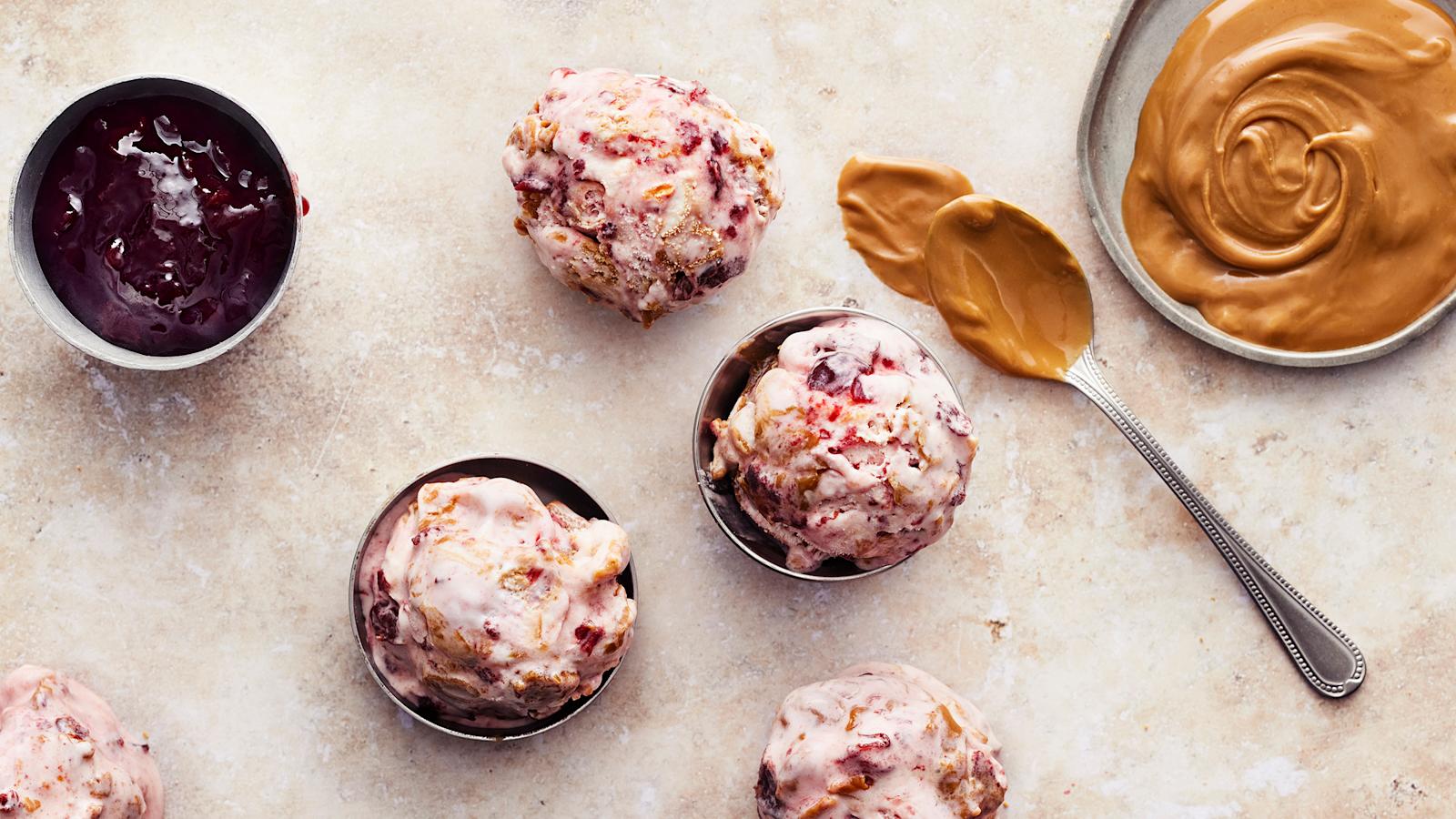 Viisi palloa vegaanista keksilevite-kirsikkajäätelöä. Palloista kaksi kulhossa ja loput pöytätason päällä. Keksilevitettä lautasella ja kirsikkahilloa kulhossa. Pöydällä keksilevitteessä käytetty lusikka.