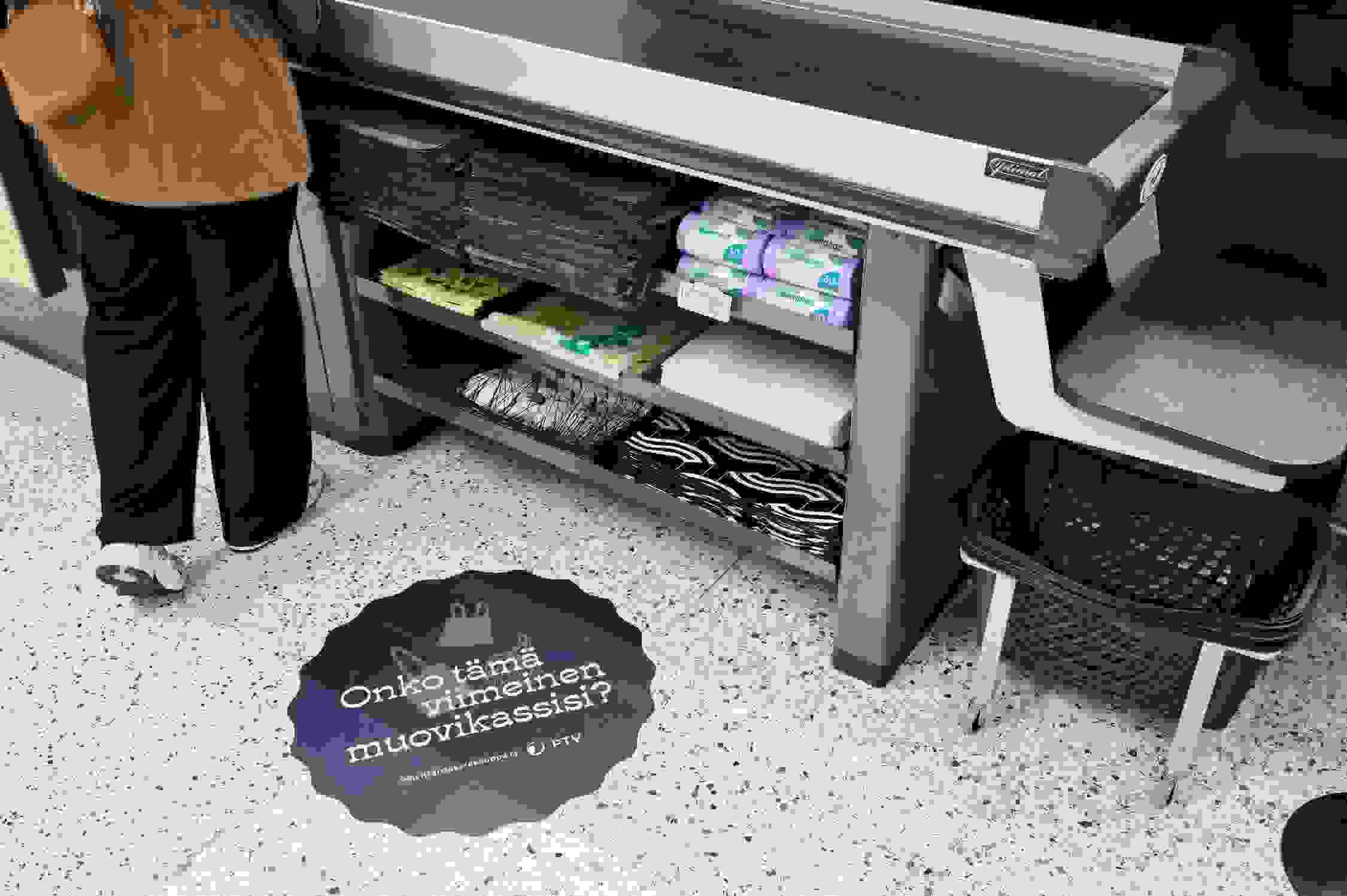 Kassalinjasto myymälässä. Kassalinjaston alla näkyy erilaisia kasseja, lattiassa Onko tämä viimeinen muovikassisi -tarra ja sen vieressä näkyy hieman asiakkaan jalkoja.