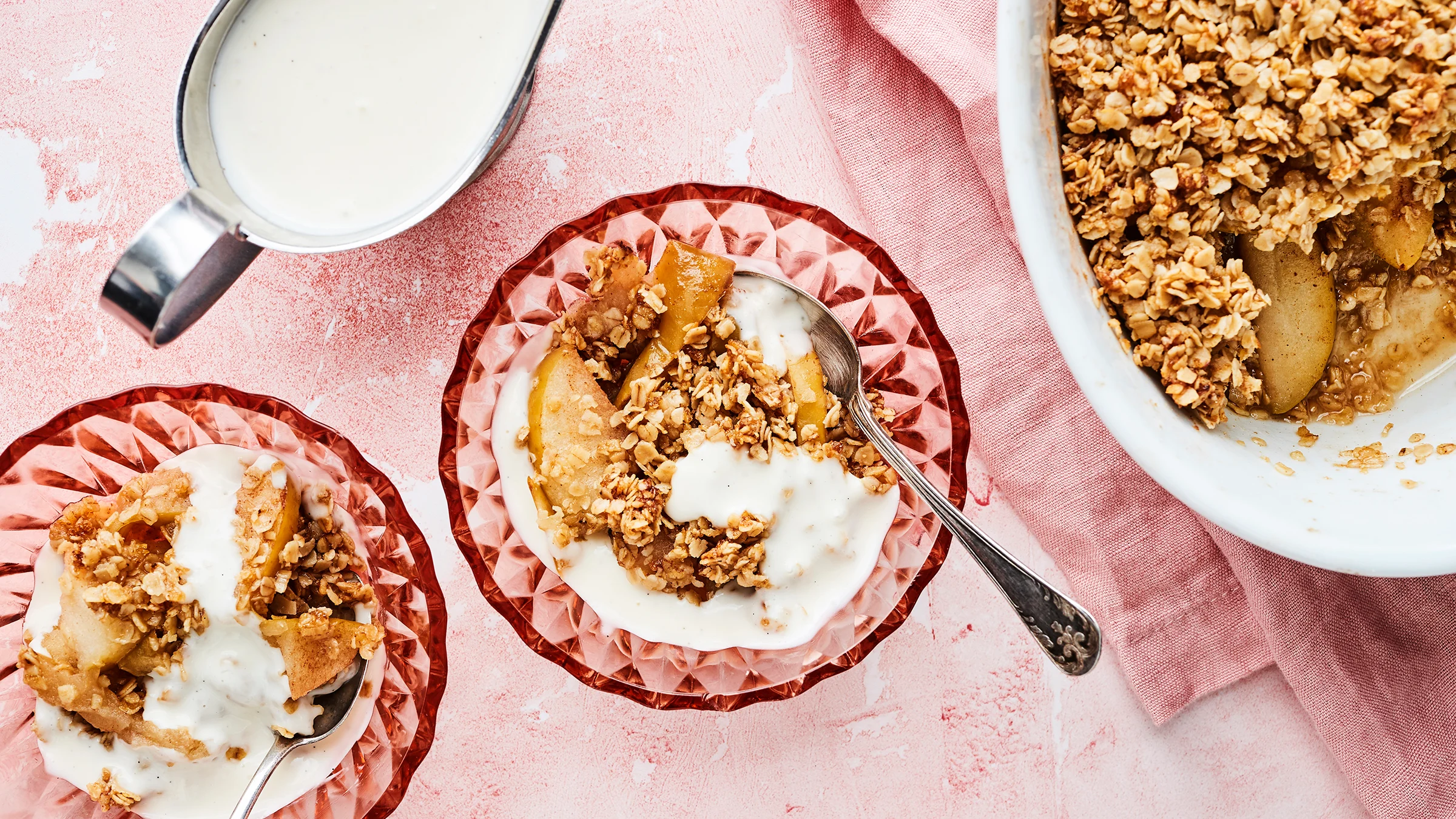 Kaura-omenapaistosta kahdessa lasikulhossa ja uunivuoassa. Uunivuoan alla vaaleanpunainen liina. Vanijakastiketta kastikekannussa.