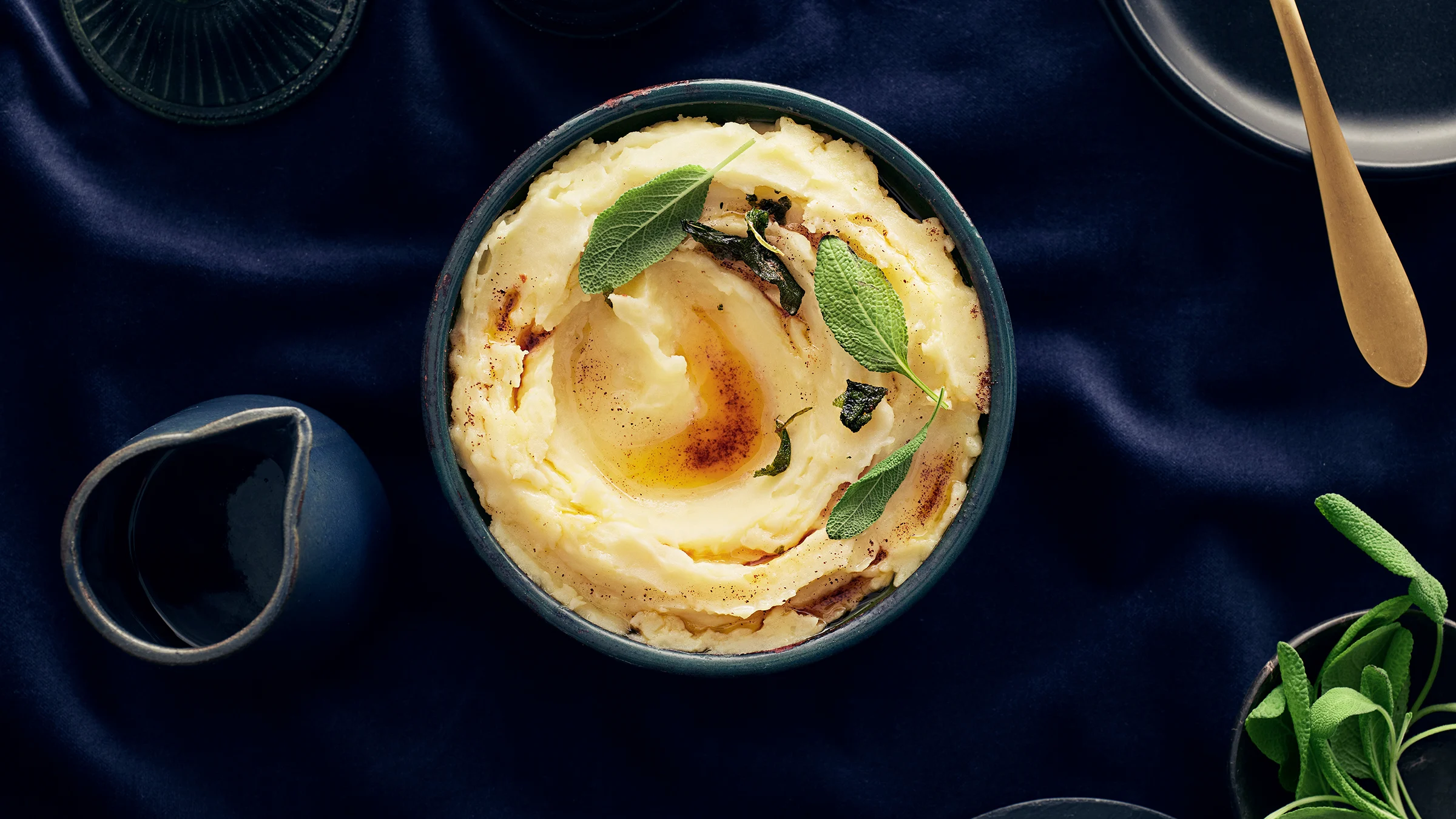 Kulhossa perunamuusia, jonka päällä salviavoita ja salvianlehtiä. Vieressä kastikekannu, jossa salviavoita.