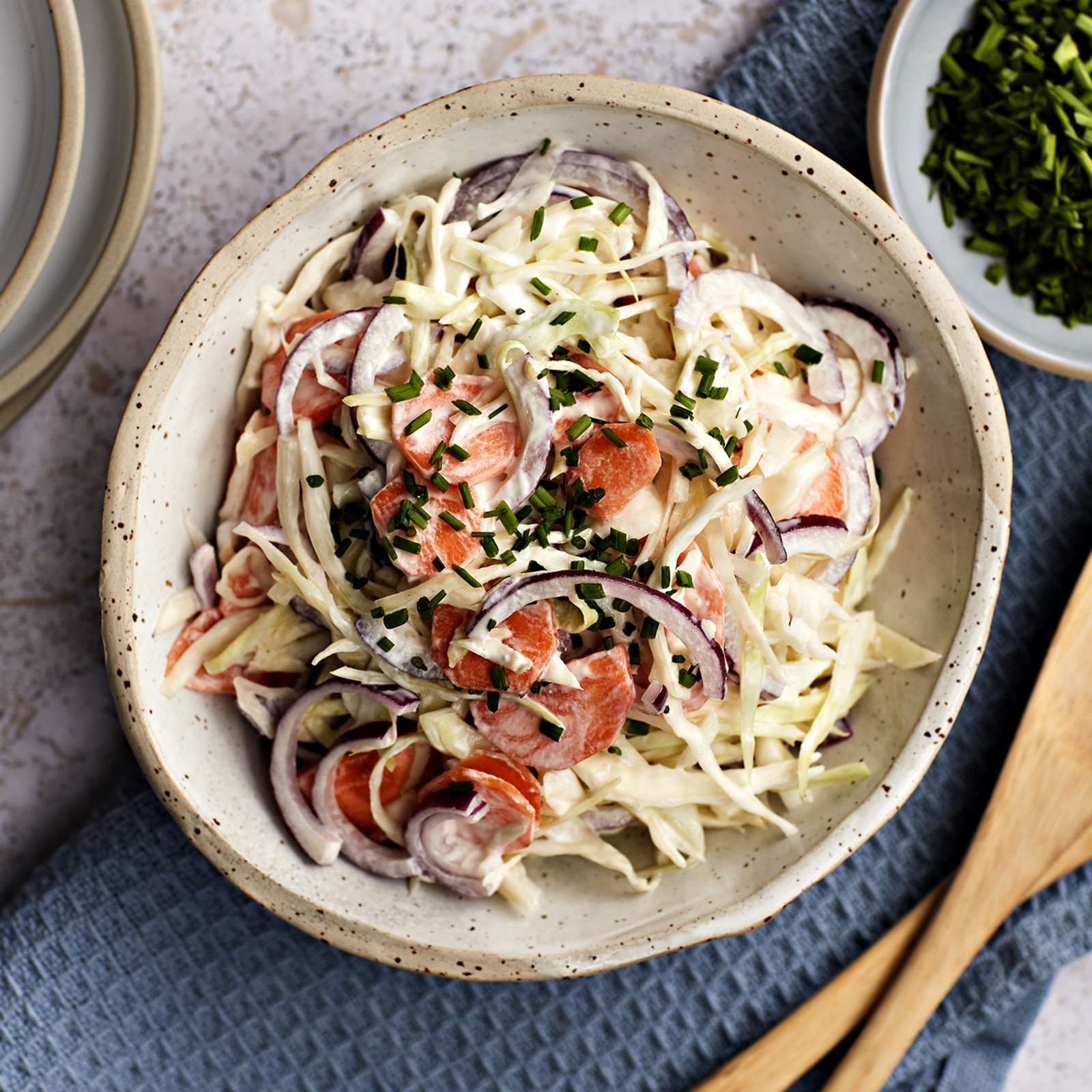 Coleslaw salaattia kulhossa, jonka alla sininen liina. Vieressä puiset salaattiottimet, ruohosipulisilppua kulhossa ja lautaspino, jonka päällä haarukoita.