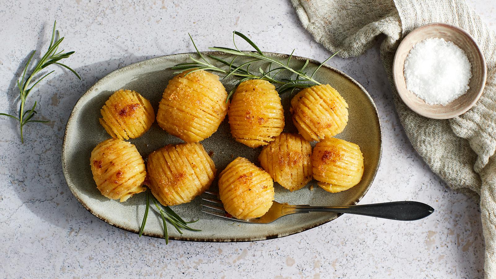 Hasselbackan perunat lautasella, jossa haarukka. Vieressä rosmariinin oksia, liina ja pieni kipollinen sormisuolaa.