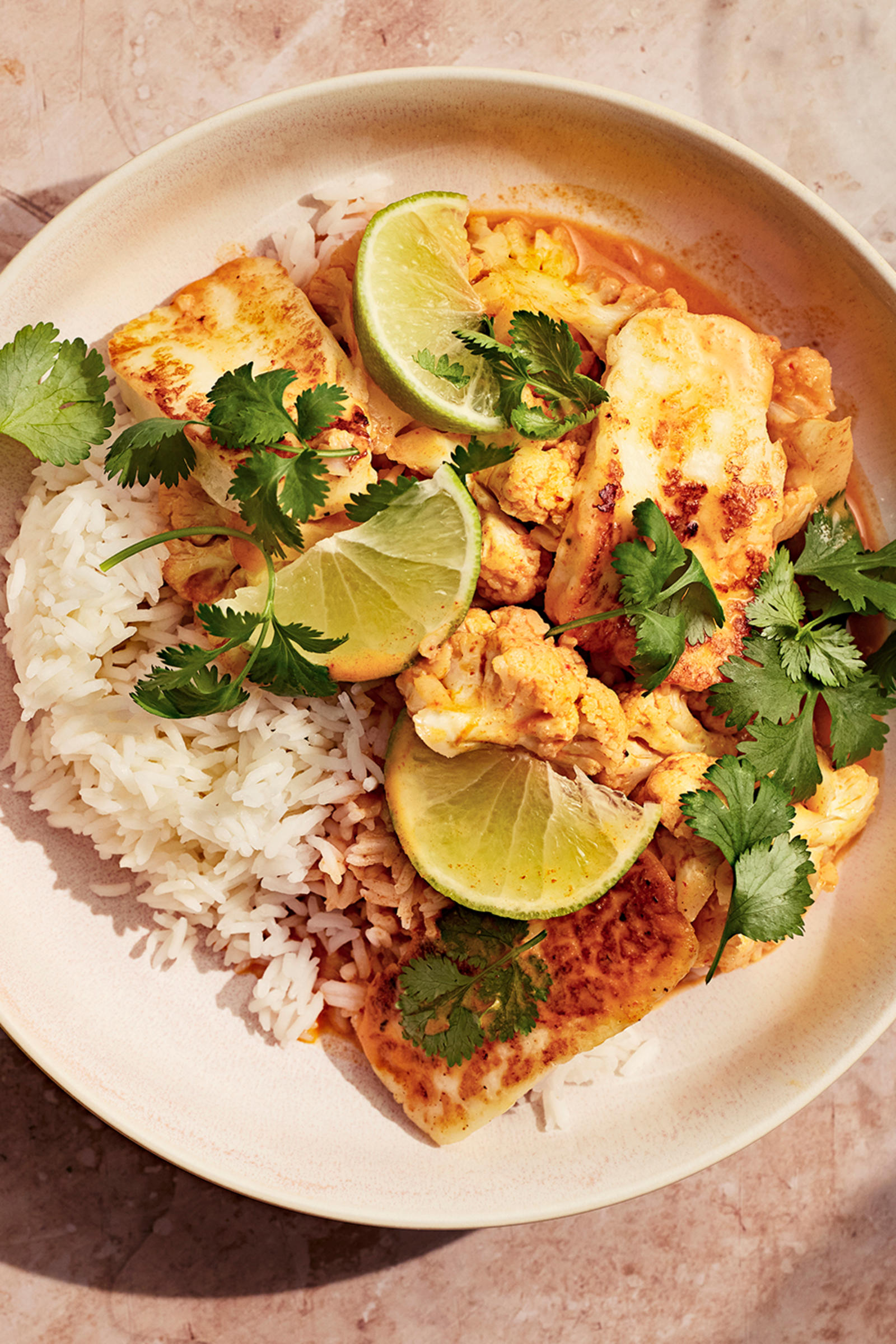 Halloumicurry ja riisiä limellä ja korianterilla koristeltuna. Annoksen vieressä vesilaseja, limejä ja beige liina.