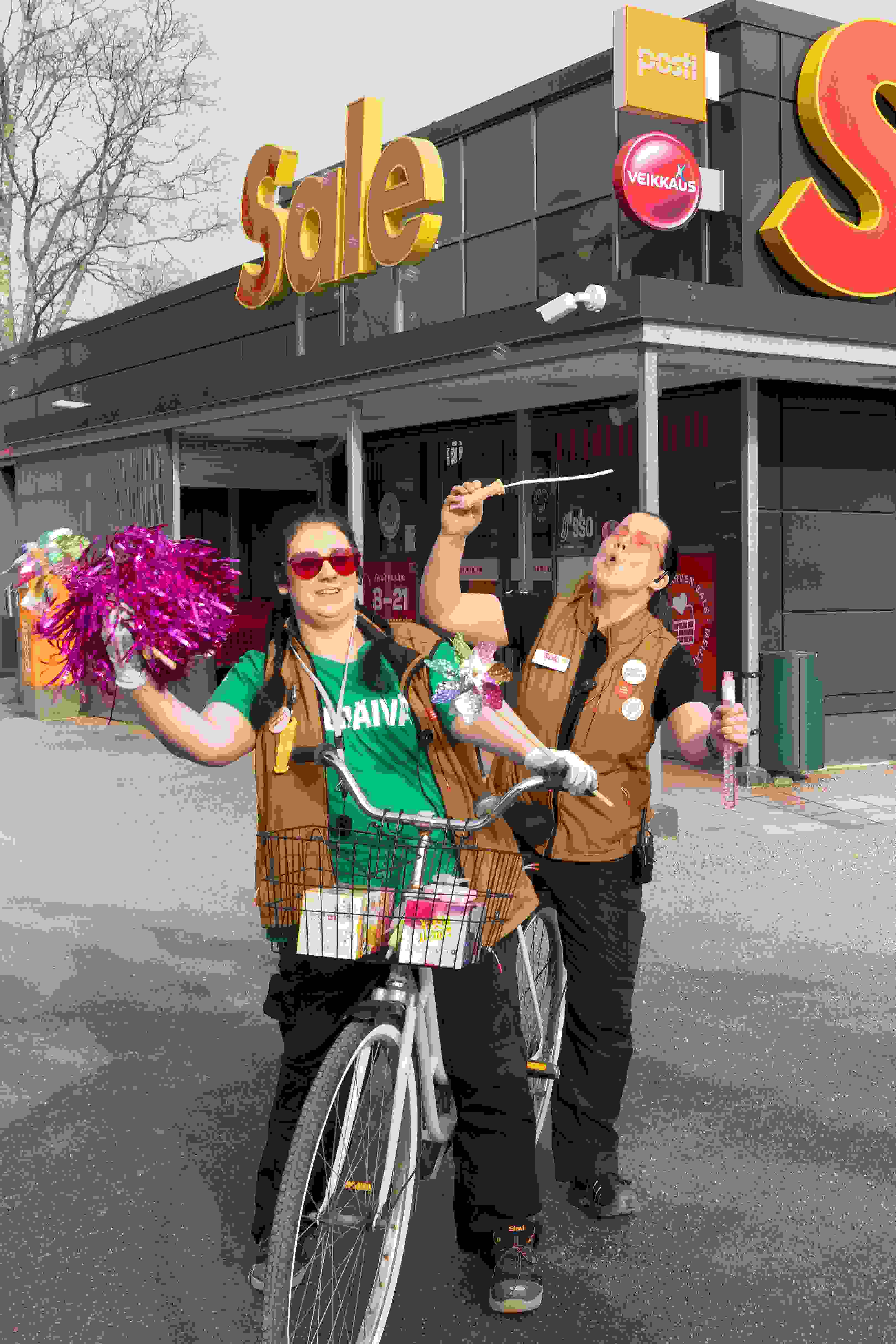 Kaksi iloista Sale-työntekijää Sale-myymälän edessä Suomusjärvellä. Heillä on pyörä sekä vappukoristeita..