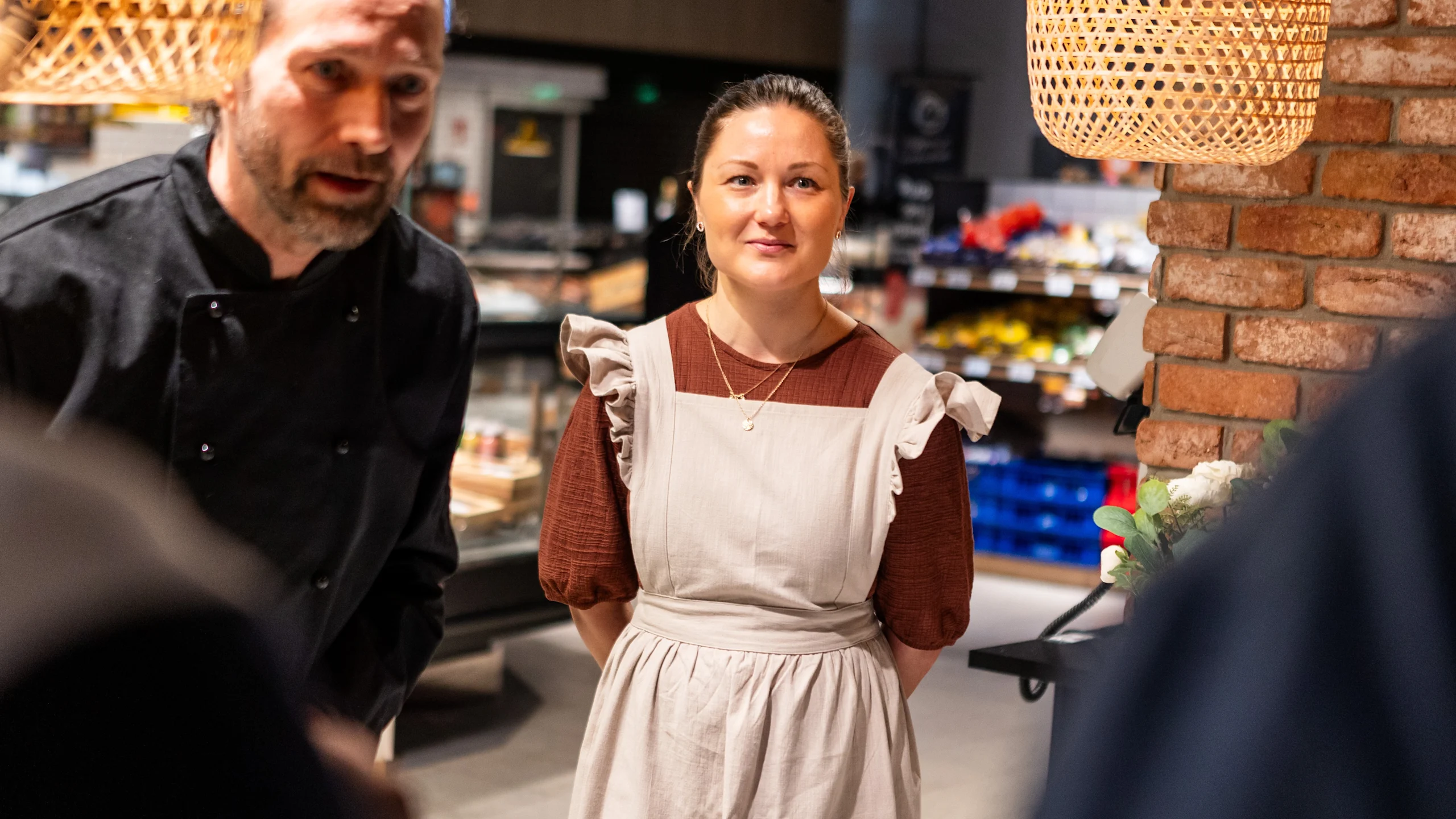 Jarmo Lipasti puhuu asiakkaille. Silpa Kinnunen seuraa vieressä kädet selän takana. Taustalla näkyy S-marketin palvelutiski sekä punainen tiiliseinä.