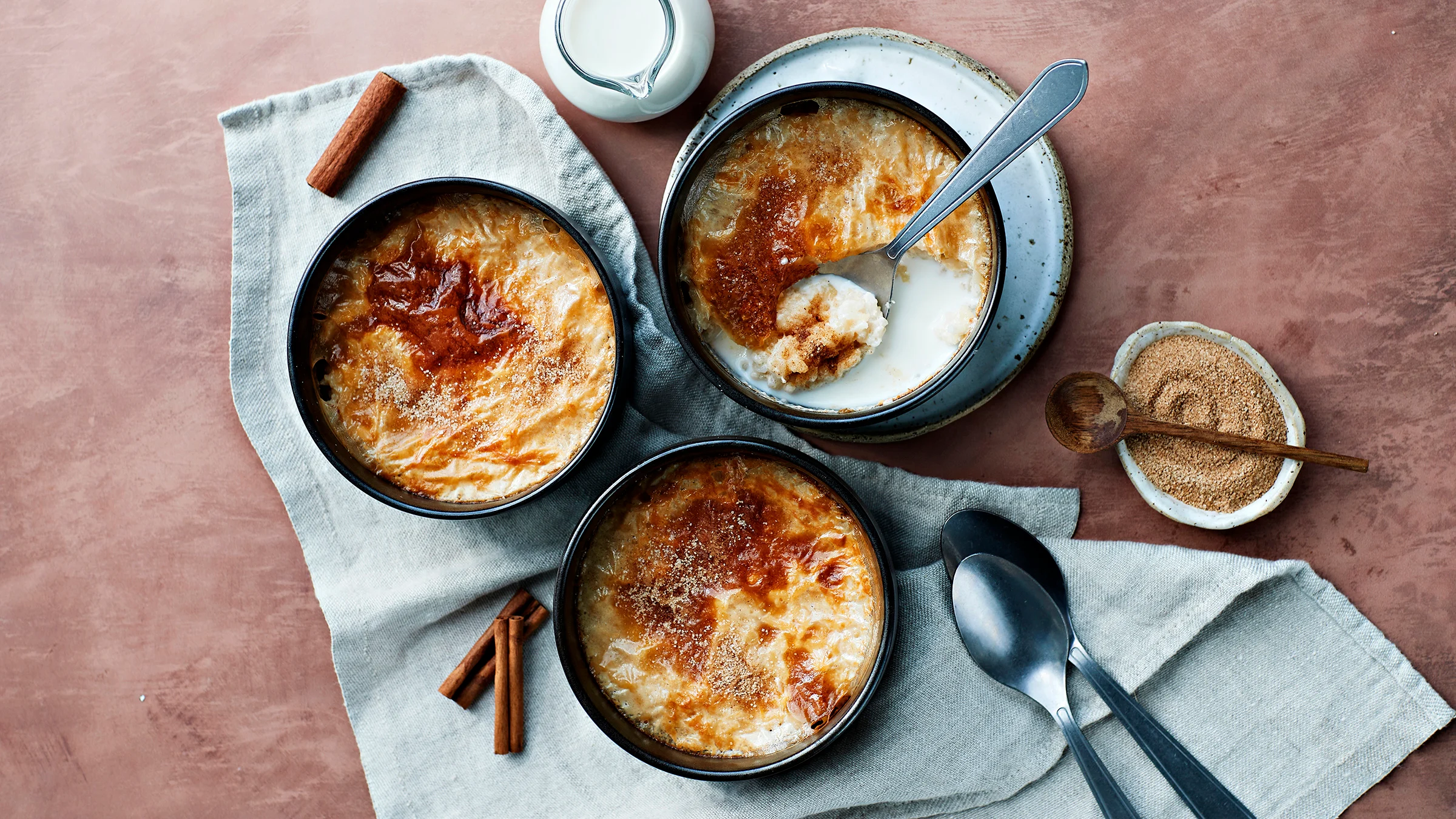 Uuniriisipuuro valmistettuna kolmessa kulhossa. Kulhojen alla liina ja vieressä lusikoita, kanelisokeria ja maitokannu.