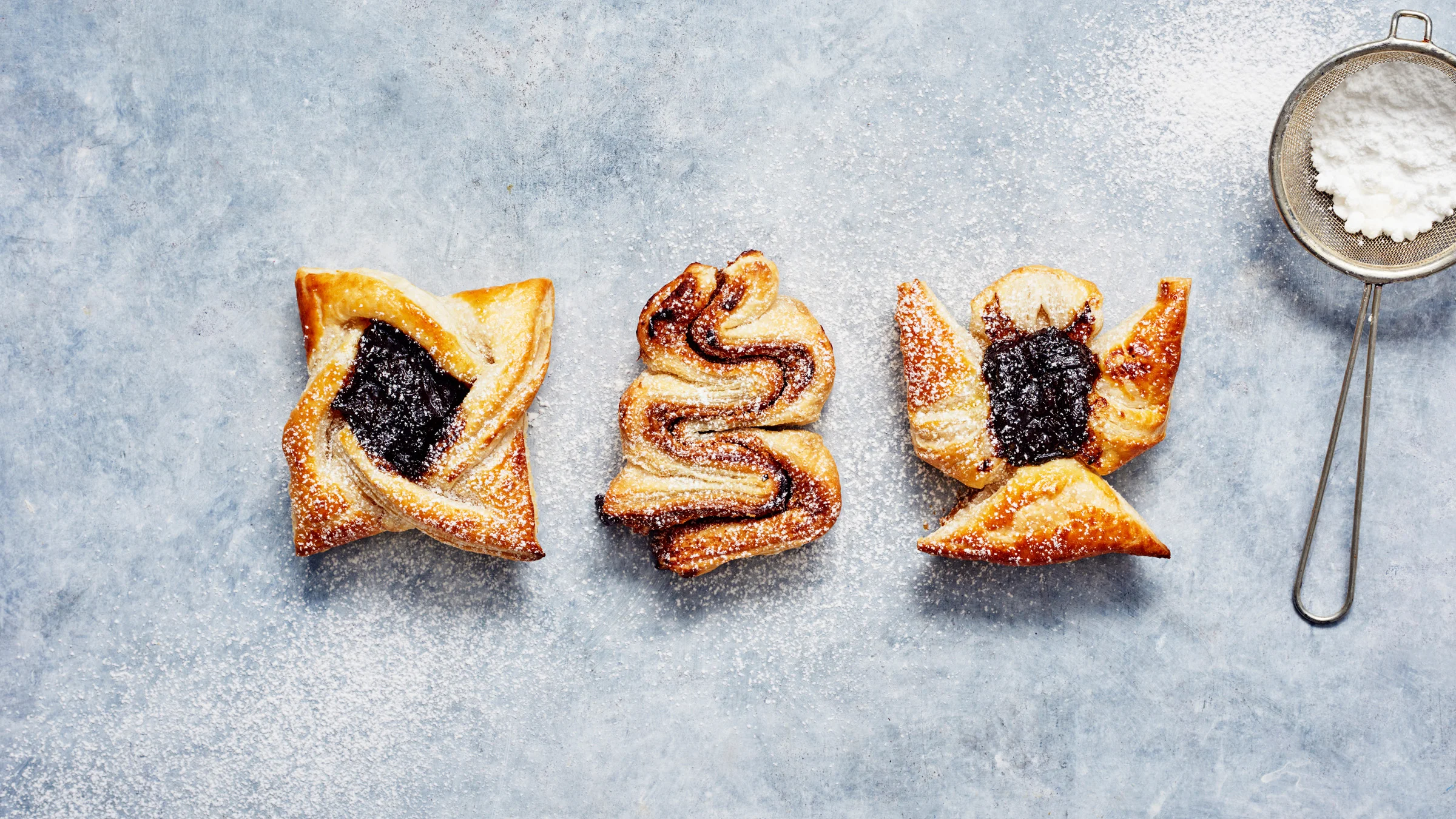 Harmaalla taustalla kolme joulutorttua: timanttitorttu, kuusen muotoinen torttu ja enkelitorttu. Vieressä pieni siivilä, jossa tomusokeria.