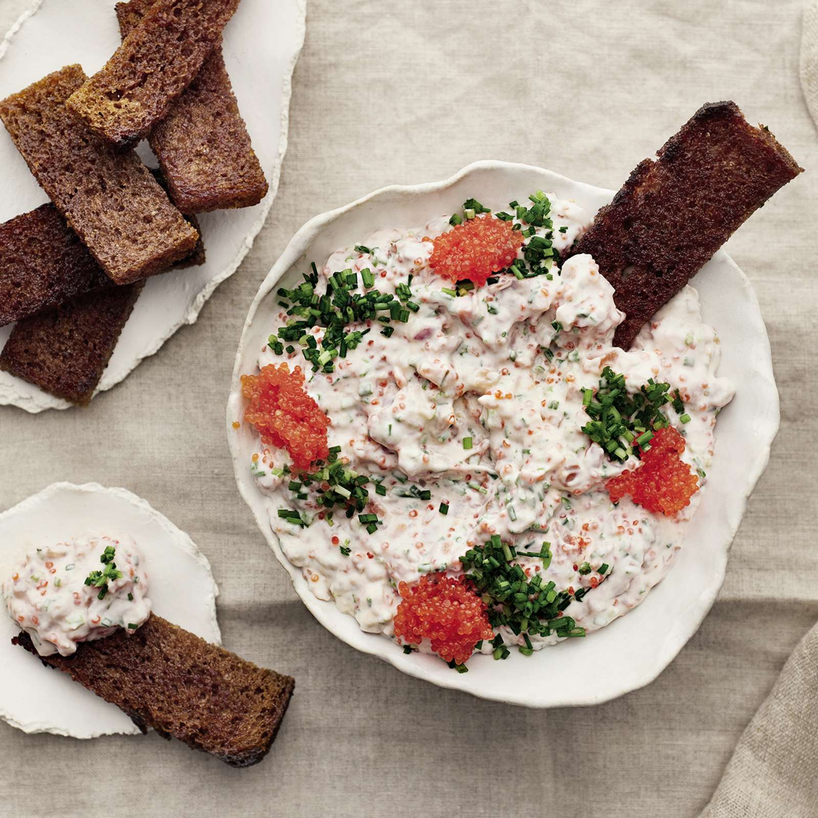 Vegaanista skagenia kulhossa merilevävalmisteella ja ruohosipulilla koristeltuna. Kulhoon dipattuna yksi leipätikku. Lisää leipätikkuja lautasilla kulhon vierellä.