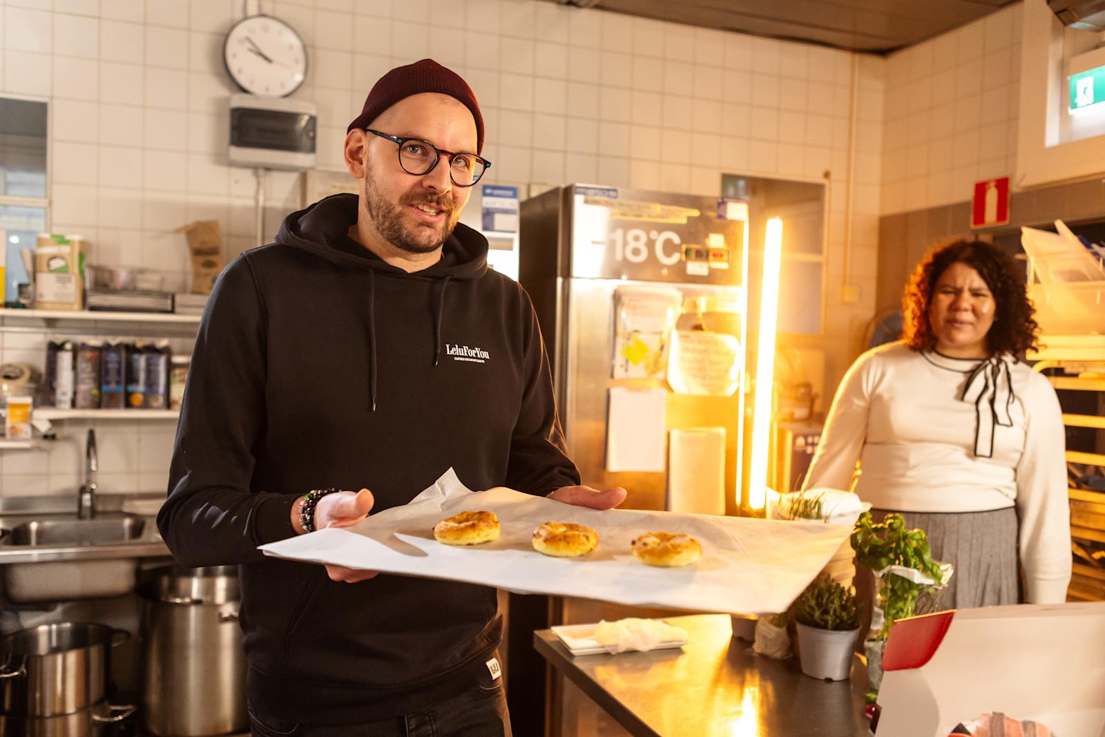 Etualalla Olli-Pekka Ojansivu pitelee uunipeltiä, jonka päällä on leivonnaisia. Taustalla Julia Lounas katsoo tarkkaavaisesti.