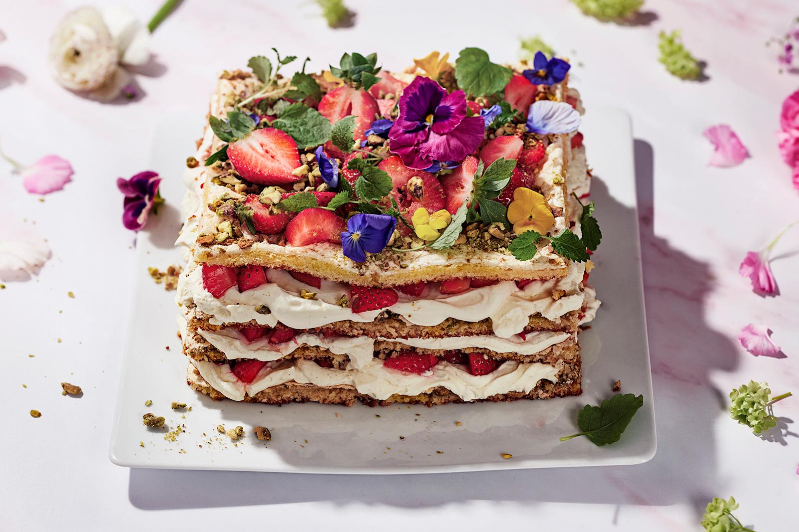 Pistaasi-britakakku, joka on koristelut orvokeilla, mansikoilla ja mintunlehdillä. Kakun ympärillä vaaleanpunaisia kukkia ja kukkien terälehtiä.