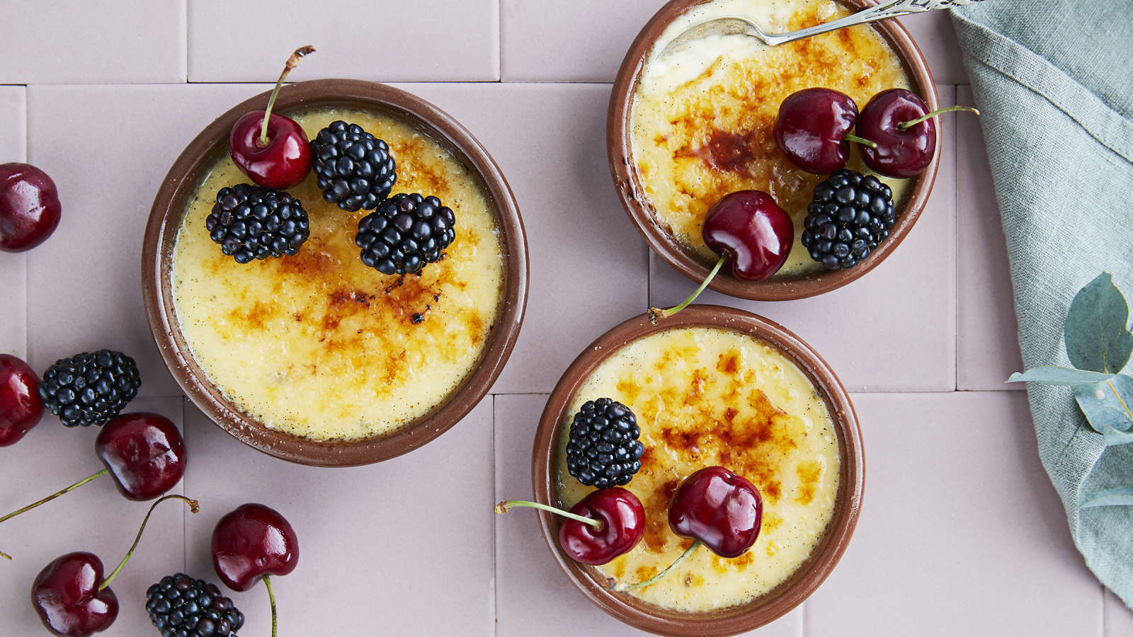 Kolme crème brûléeta, jotka on koristeltu tuoreilla kirsikoilla ja karhunvadelmilla. Kulhojen veressä liina sekä koristeluun käytettyjä marjoja.
