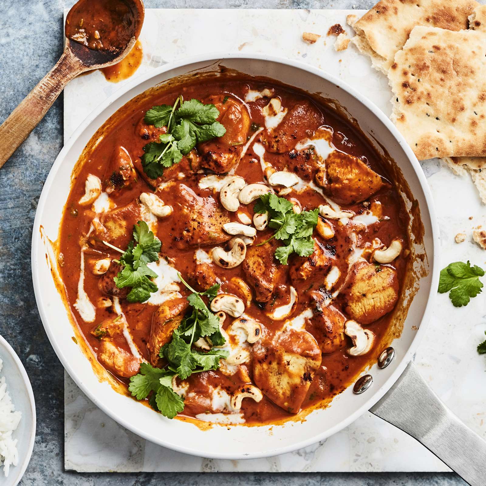 Butter chicken teräksisessä paistinpannussa. Vieressä puinen kauha, murrettua naanleipää, korianteria ja kulhollinen riisiä.