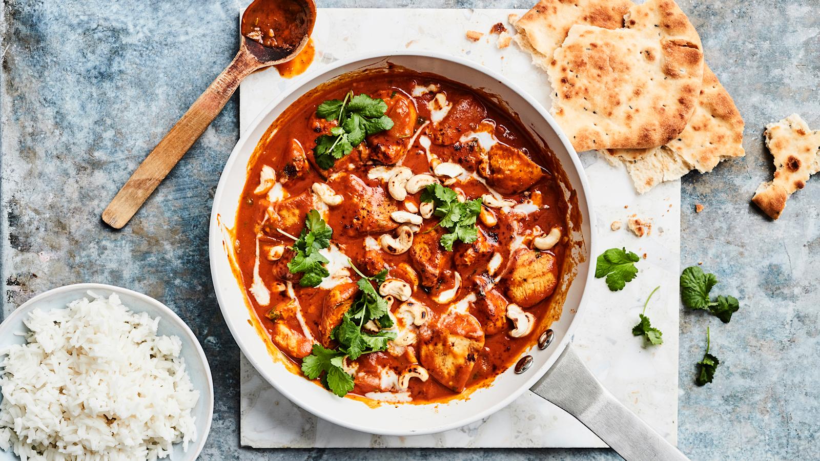 Butter chicken teräksisessä paistinpannussa. Vieressä puinen kauha, murrettua naanleipää, korianteria ja kulhollinen riisiä.