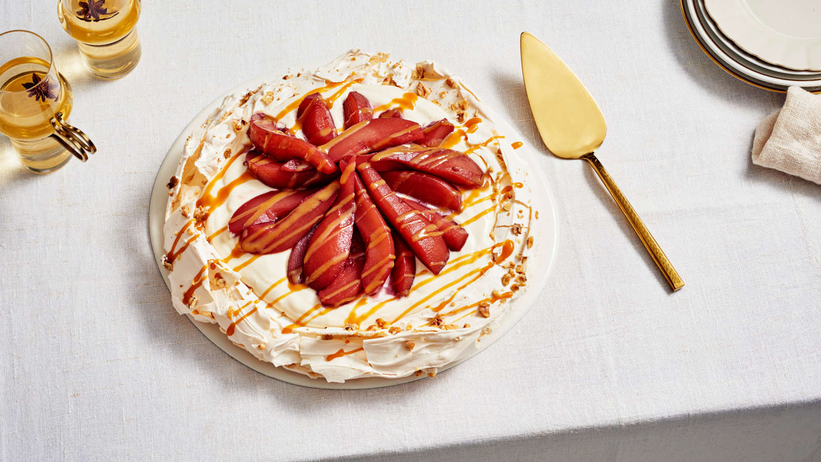 Glögimarinoiduilla päärynöillä ja kinuskikastikkeella täytetty pavlova tarjottimella. Vieressä kultainen kakkulapio ja kaksi lasia vaaleaa glögiä.