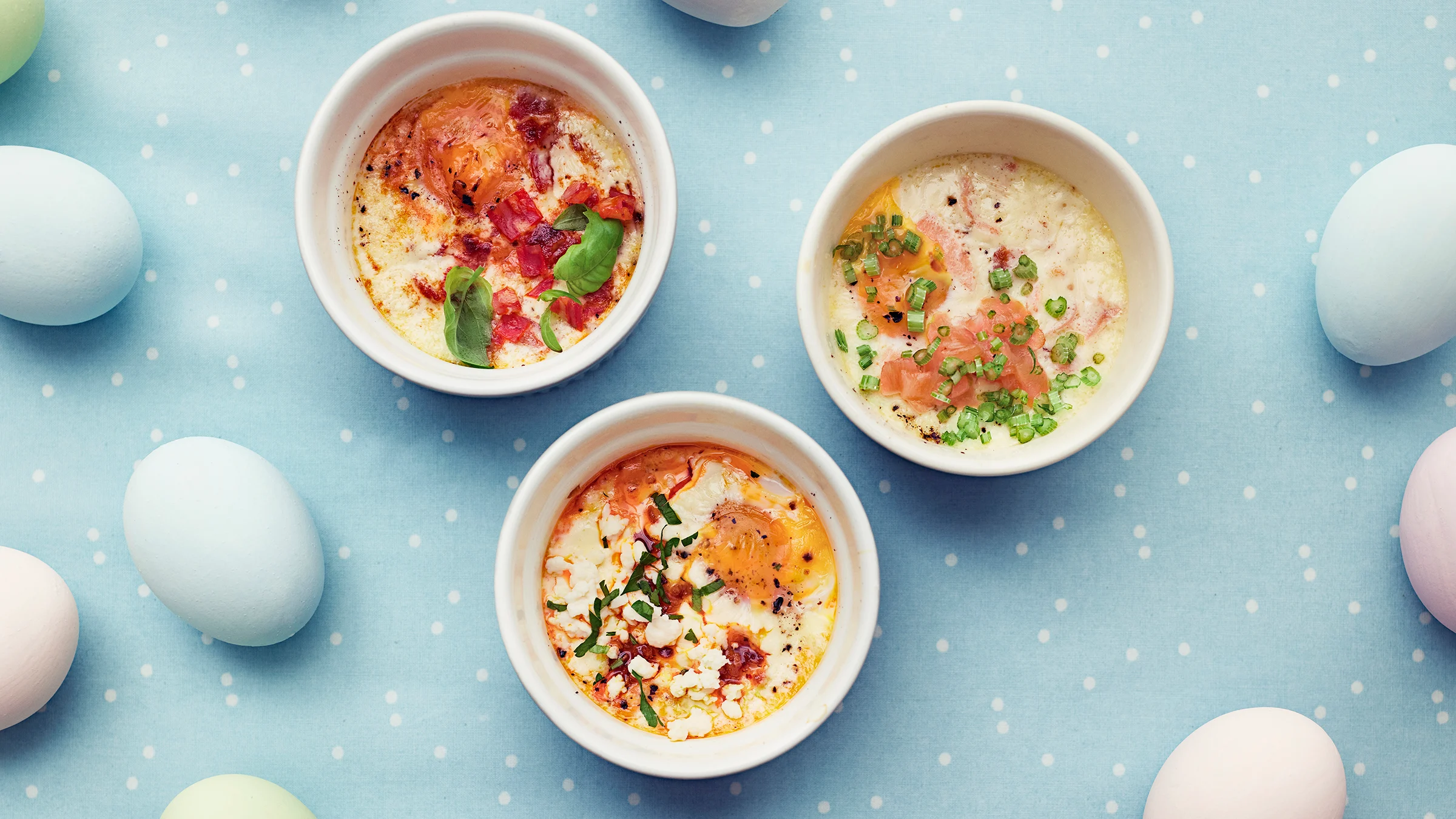 Uunimunat kolmella tavalla maustettuna pienissä vuoissa. Vieressä pastellisävyin värjättyjä kananmunia.