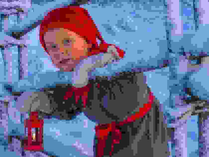 Tonttuasuun pukeutunut poika kurkkii kumisen aidan raosta ja hänellä on kädessään lyhty.