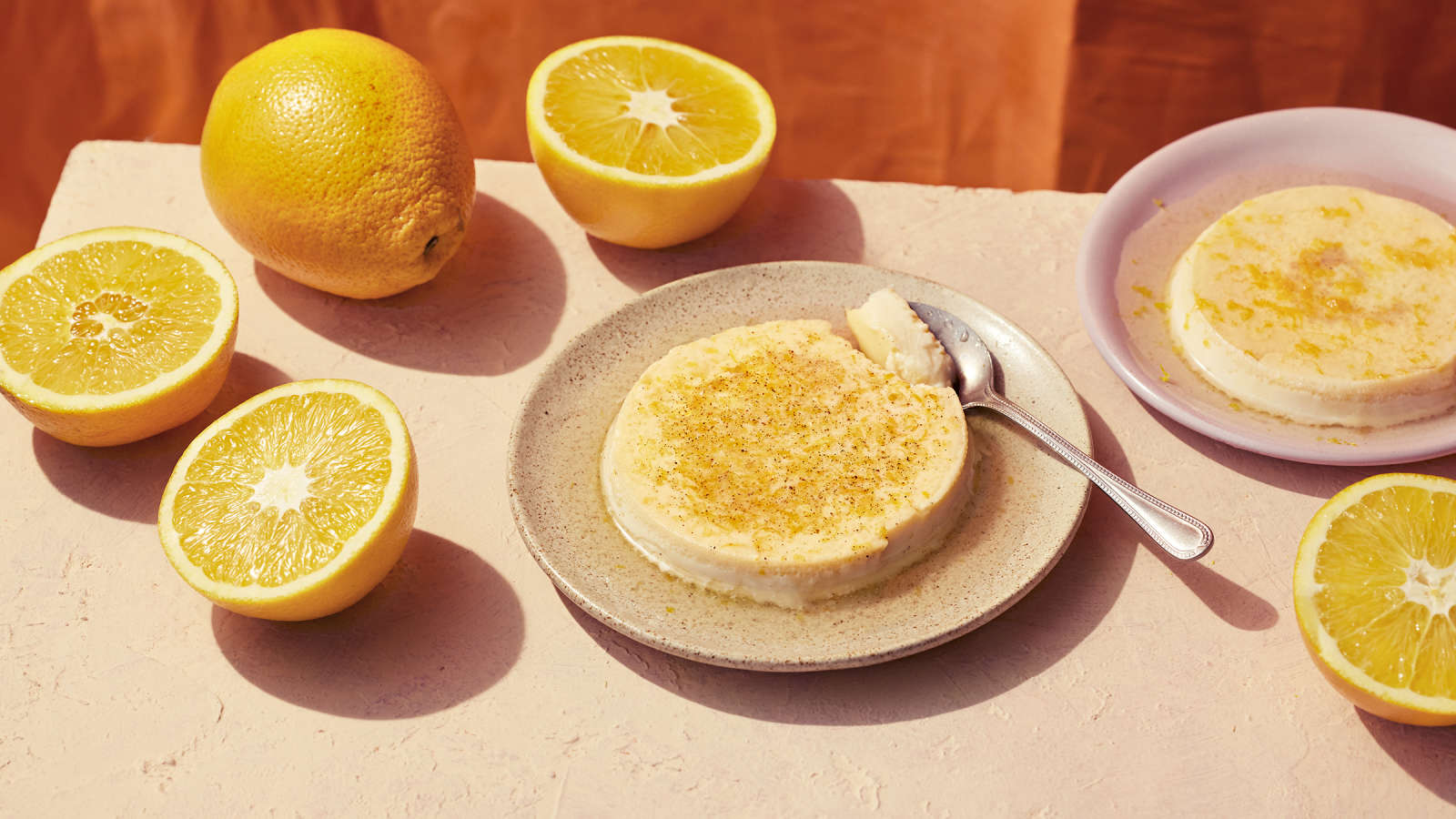 Appelsiininen flan eli paahtovanukas lautasella, jossa lusikka. Vieressä toinen vanukas ja puolitettuja appelsiineja. 