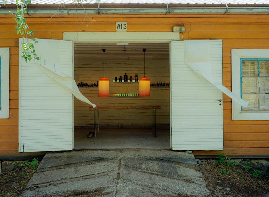 Keltaisen puutalon valkoiset pariovet ovat auki. Sisällä näkyy pöytä ja kaksi oranssia lamppua.