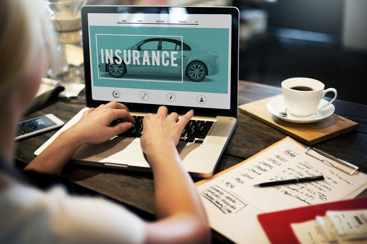 Over-shoulder view of someone working on a laptop looking for insurance online. A coffee cups sitting to the side with paperwork scattered about. Compare auto quotes