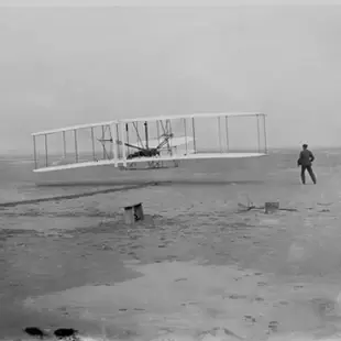 A man stands on a sandy beach near an early aircraft that is hovering just a few feet about the ground.