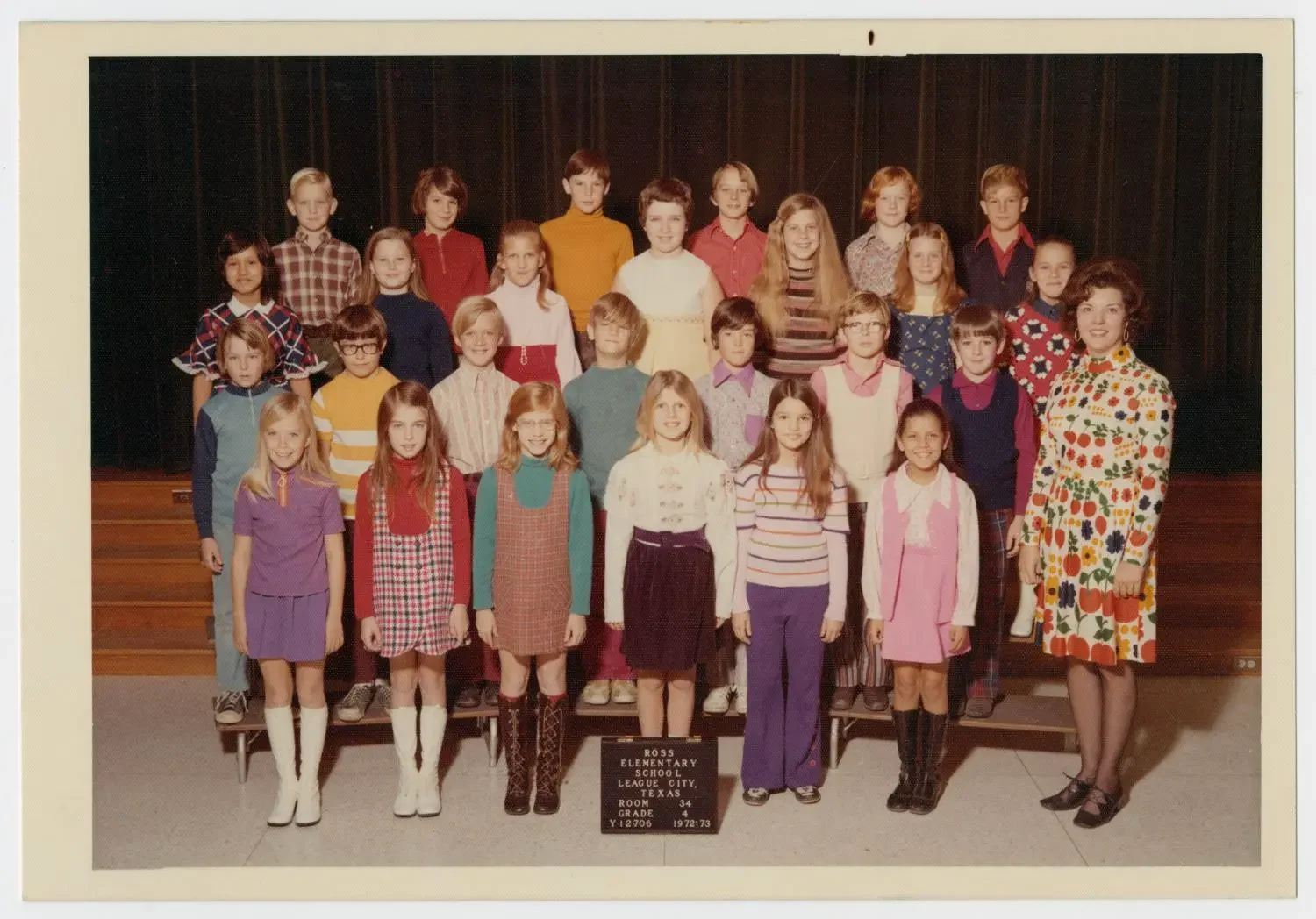US, early 1970s: The students and their teacher in this school class photo are wearing bright outfits. Girls, breaking the norm, are wearing trousers. Boys are wearing pink shirts. Notice how the girls are wearing long socks with short skirts?