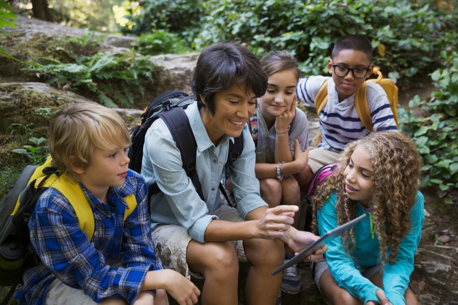 Female teacher surrounded by four children engaging with the digital content on a tablet during a field trip
