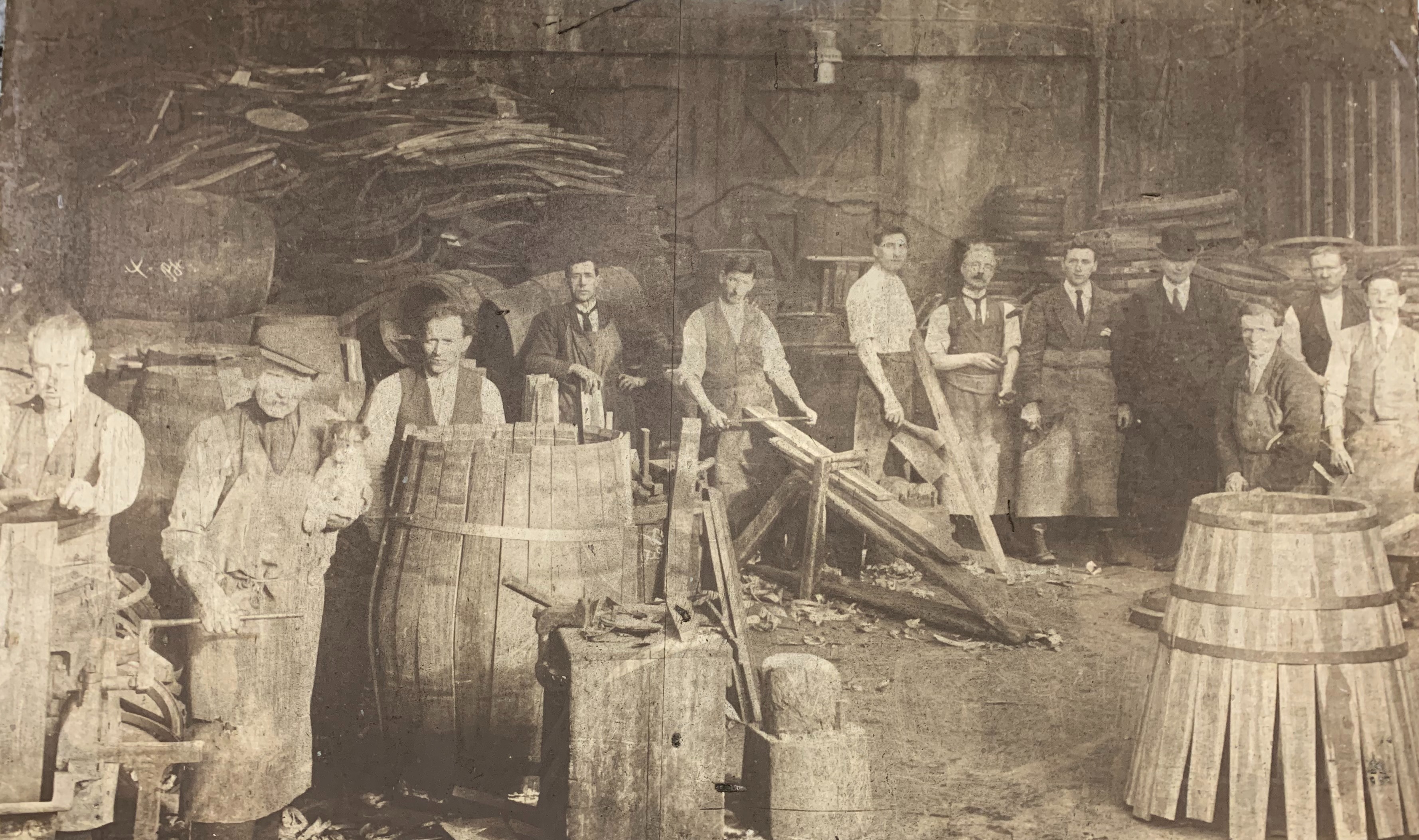 A historical photo of a group of men posing for the camera surrounded by barrels who appear to work in a whiskey distillery.