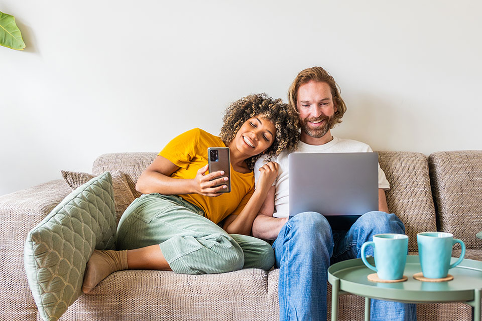 A couple looking at a phone & laptop from a sofa