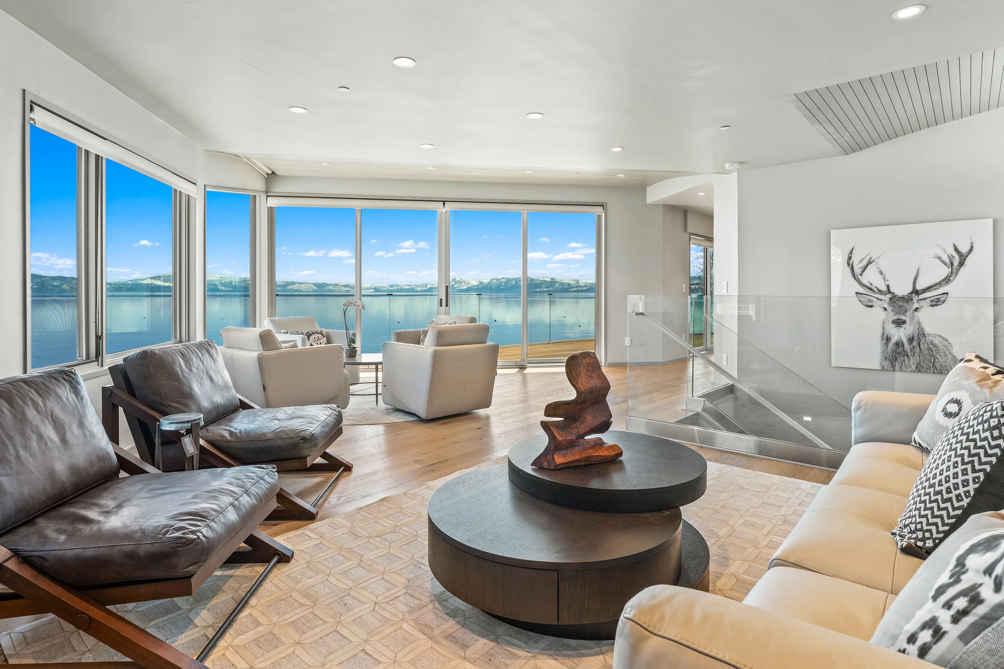 Alt text: Interior sitting area with large glass windows overlooks a lake with boats and mountains. Sunny sky.
Photo Credit: Vista Estate Visuals