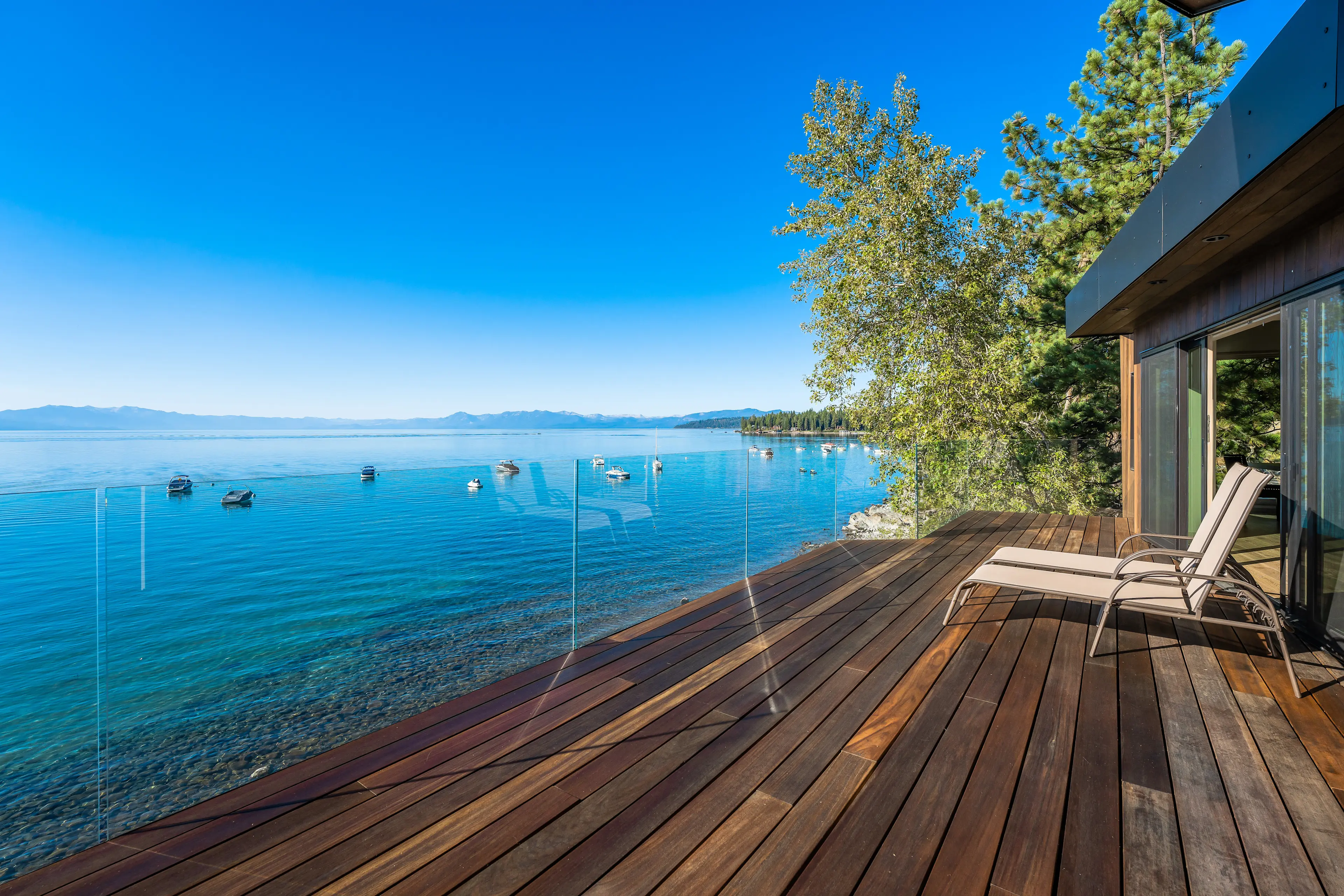Alt text: Wooden deck with lounge chairs overlooks a lake with boats and mountains. Sunny sky.
Photo Credit: Vista Estate Visuals