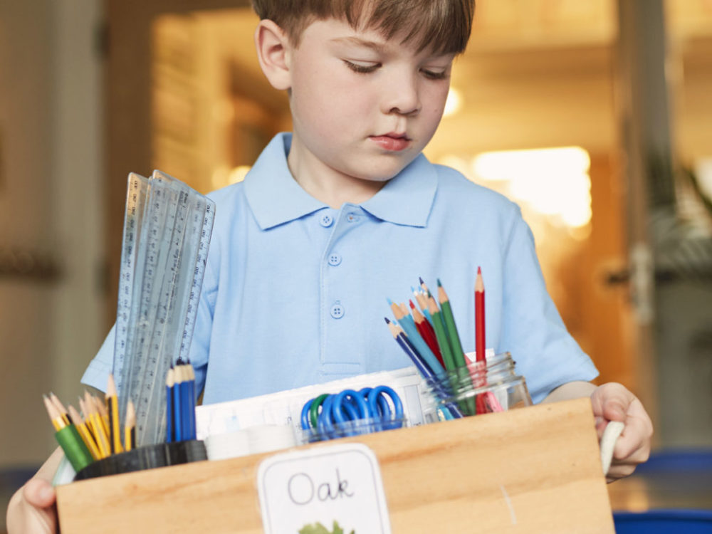 Schoolboy carrying box of pencils in classroom at primary school